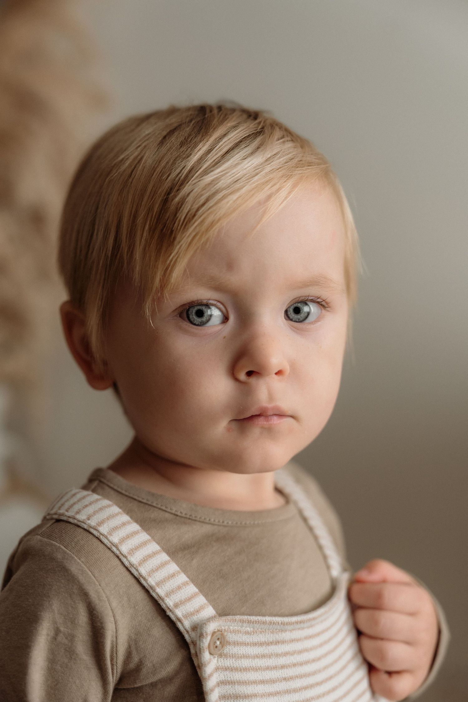A young child with bright blue eyes wearing a beige sweater and striped overalls looks directly at the camera.