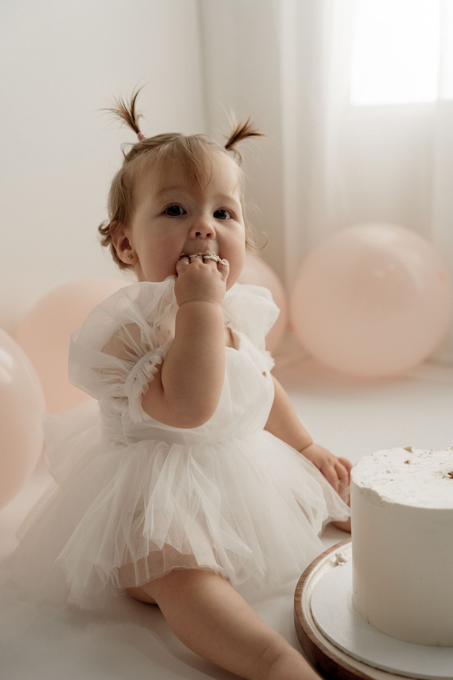 A baby in a white tulle dress plays with a cake during a first birthday photoshoot in a bright airy setting.
