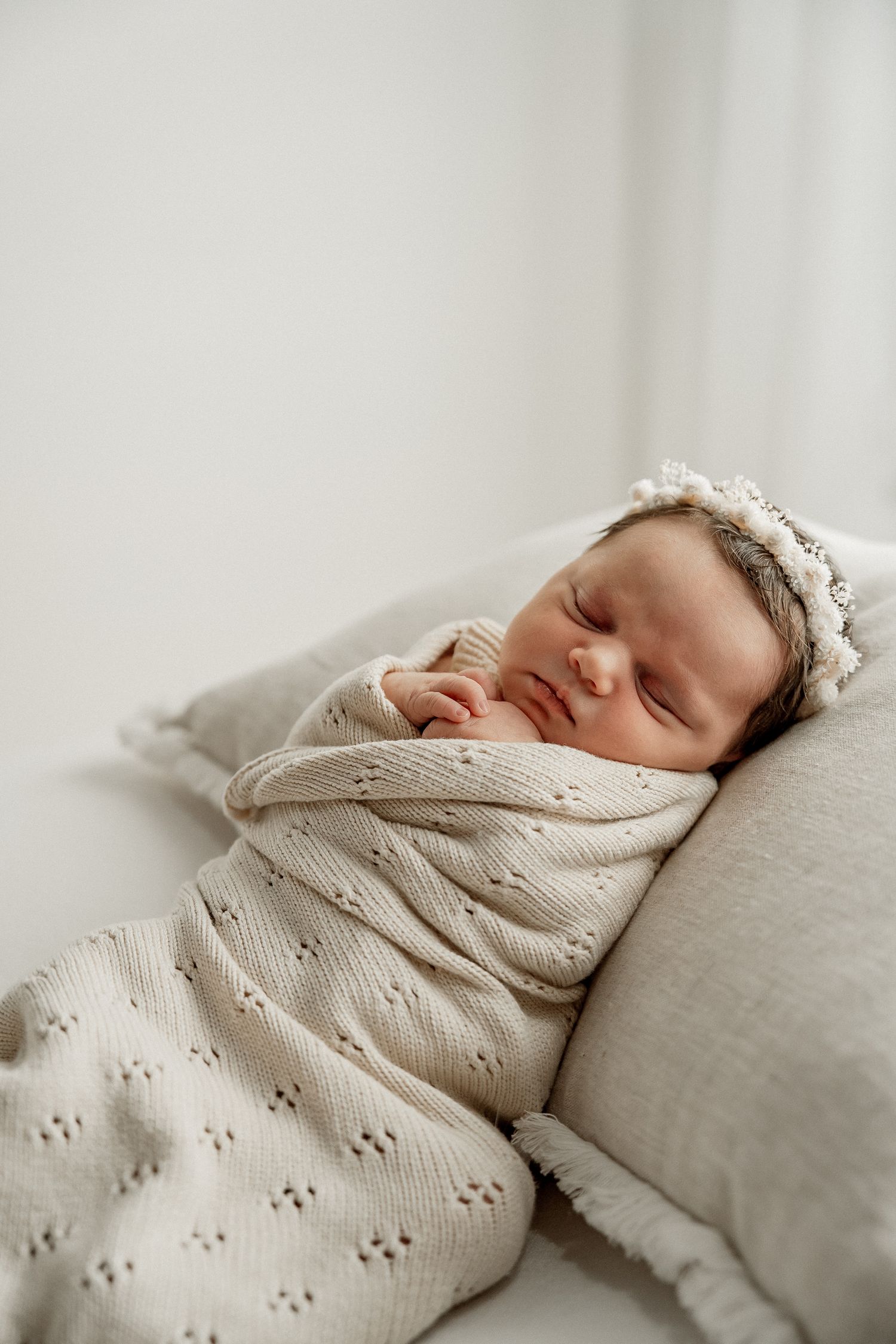 A sleeping newborn baby wrapped in a soft textured white blanket lays peacefully on light grey bedding.