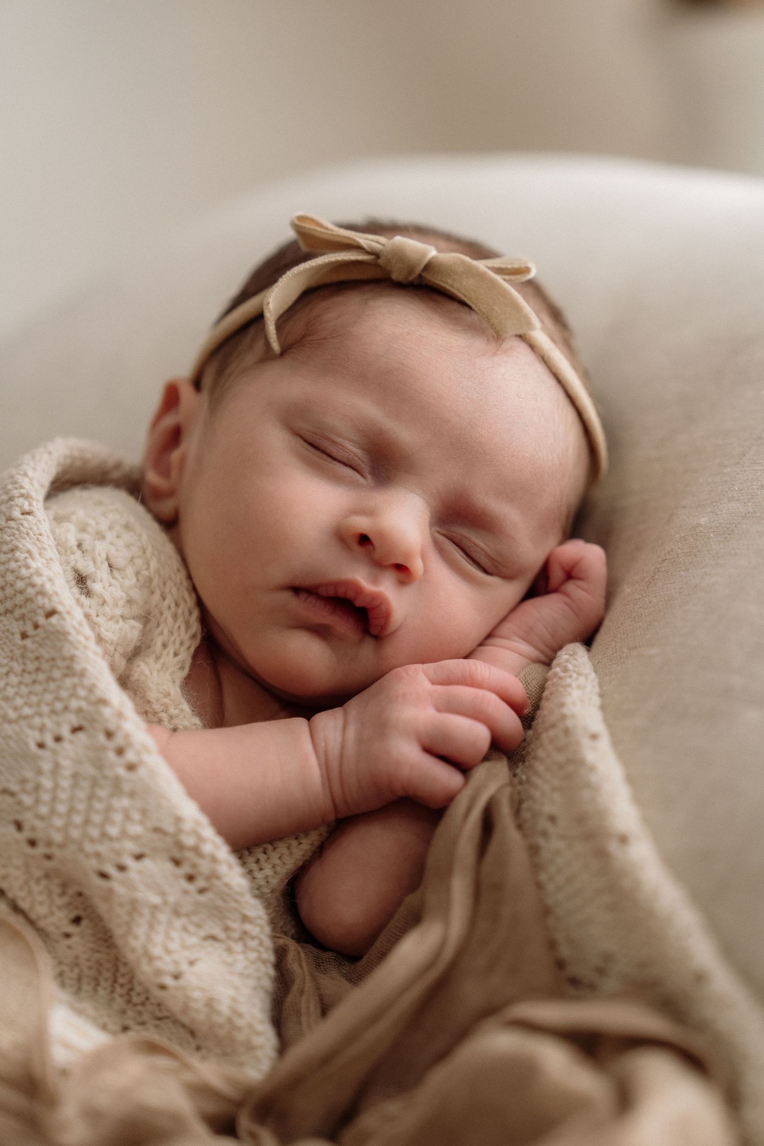Peaceful sleeping infant wrapped in beige knit blanket wearing tan bow headband on white surface.