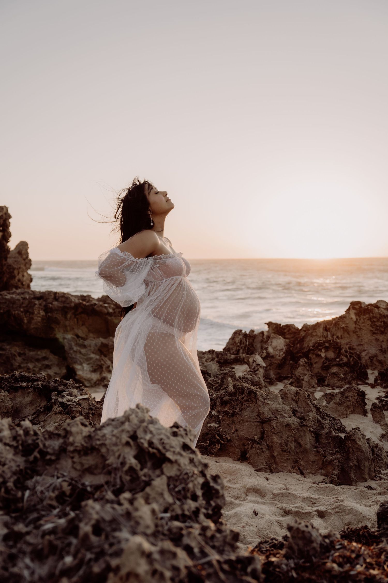 A pregnant woman in a white lace dress poses on rocky coastal cliffs at sunset by the ocean.