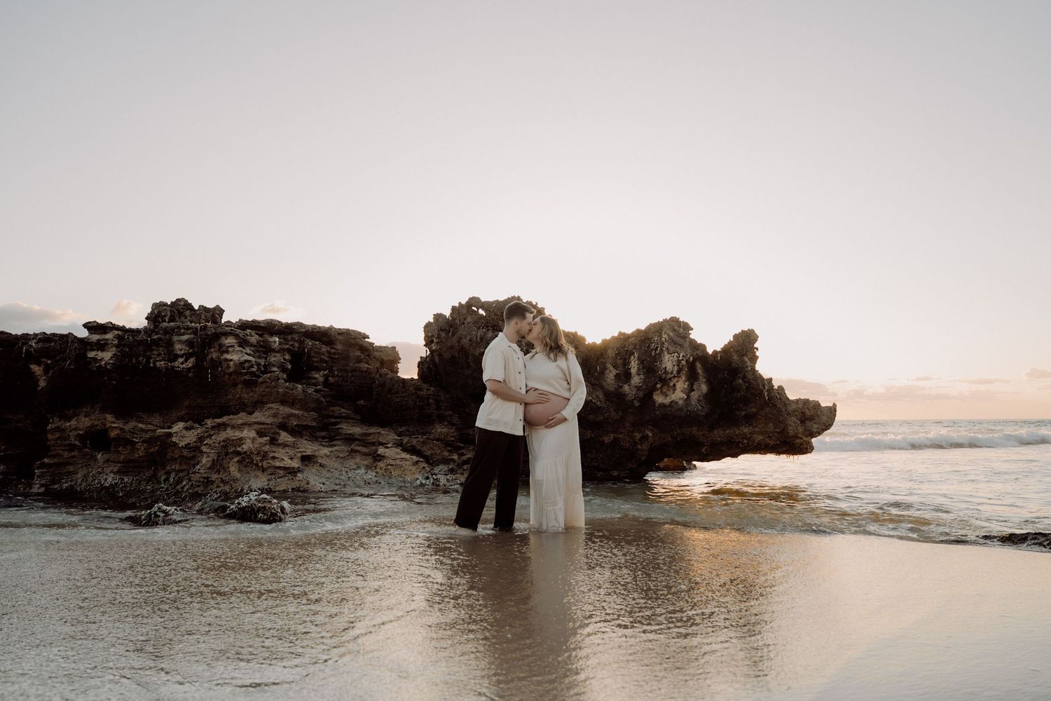 A couple embraces on a beach at sunset with dramatic rock formations and ocean waves in the background.
