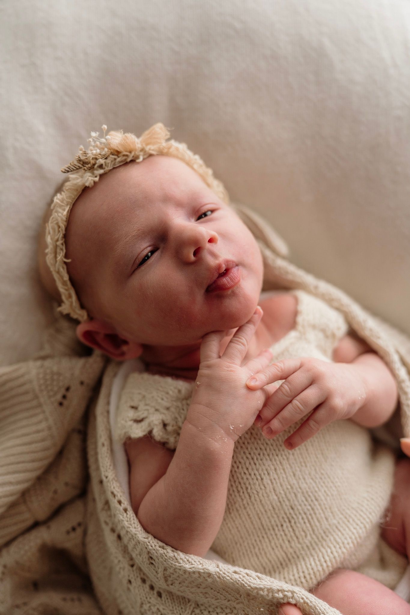 A newborn baby wearing a knit wrap rests peacefully on a soft neutral-colored backdrop during a professional photo session.