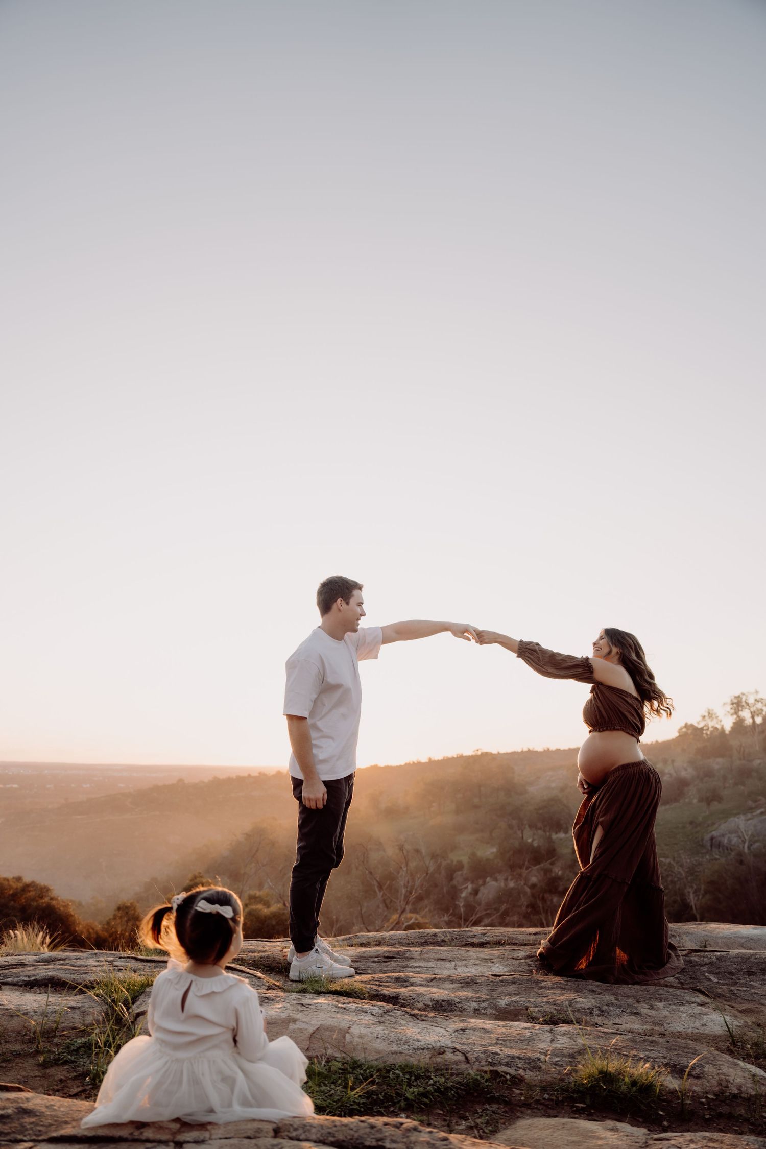 Pregnant couple holding hands at sunset on rocky overlook, with young daughter sitting nearby, golden hour maternity photo.