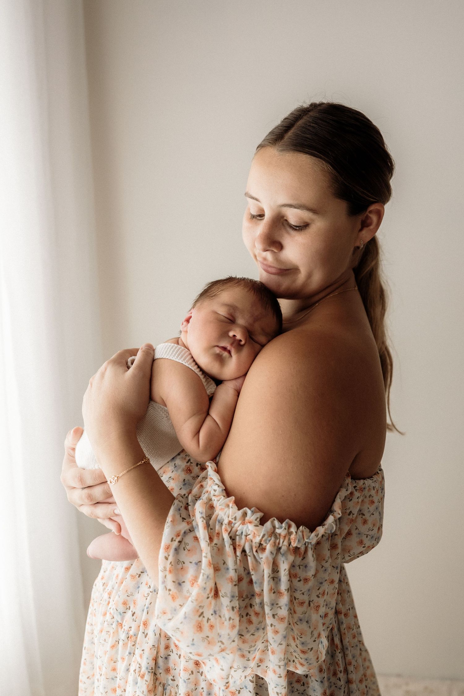 Tender moment between parent and sleeping newborn baby in soft natural window light wearing floral dress.