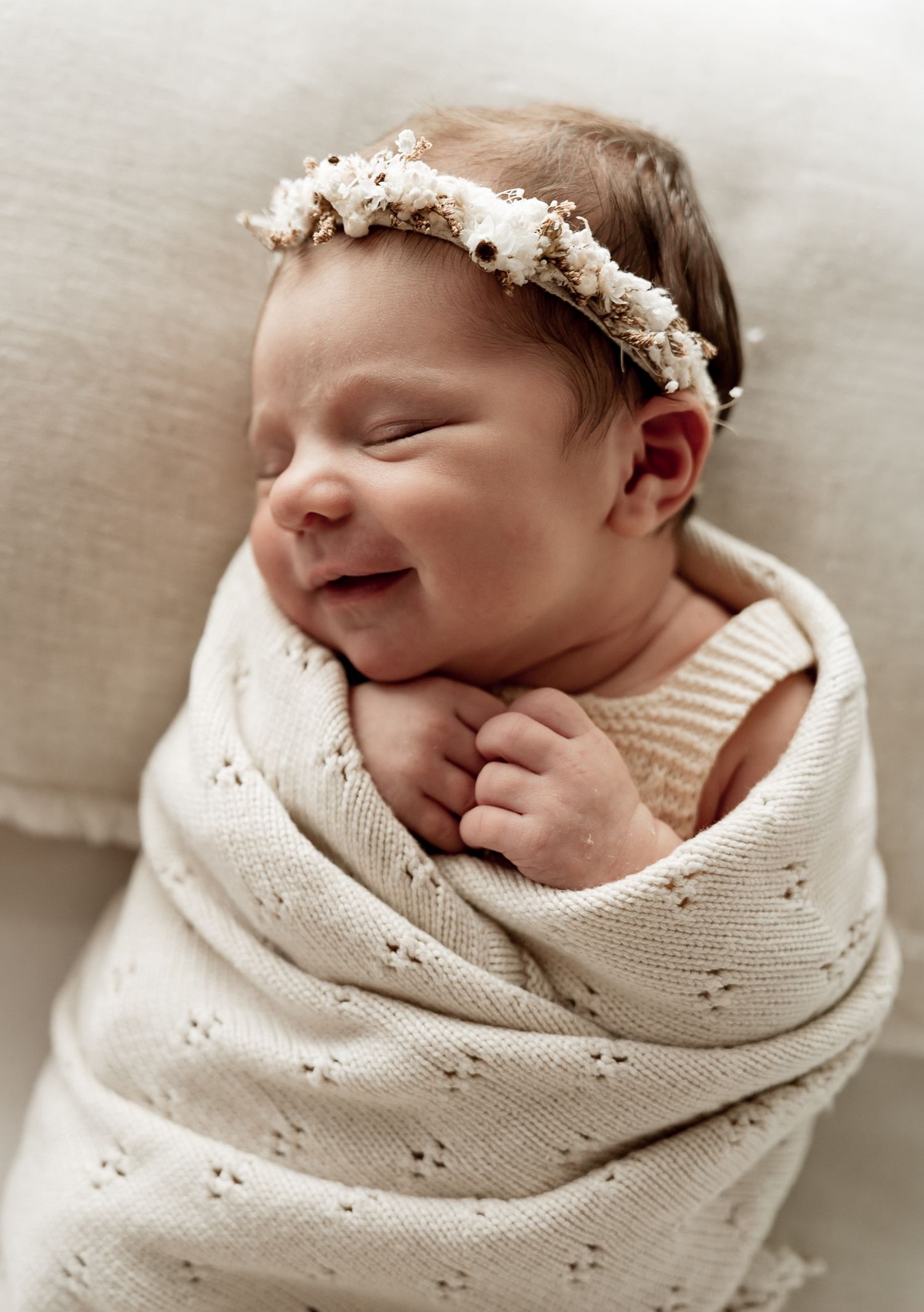 Smiling newborn baby wrapped in a cream knit blanket wearing a delicate floral headband, eyes closed peacefully.