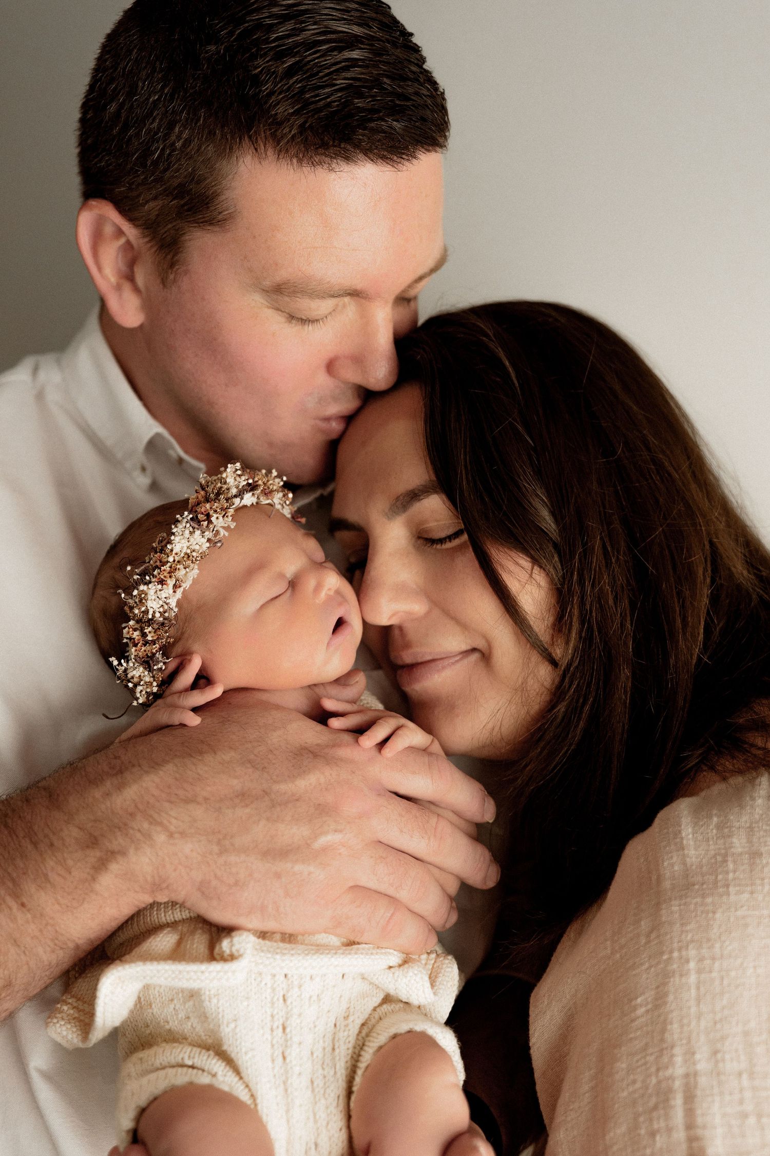 Intimate family moment showing parents lovingly embracing their newborn baby wearing a delicate floral crown.