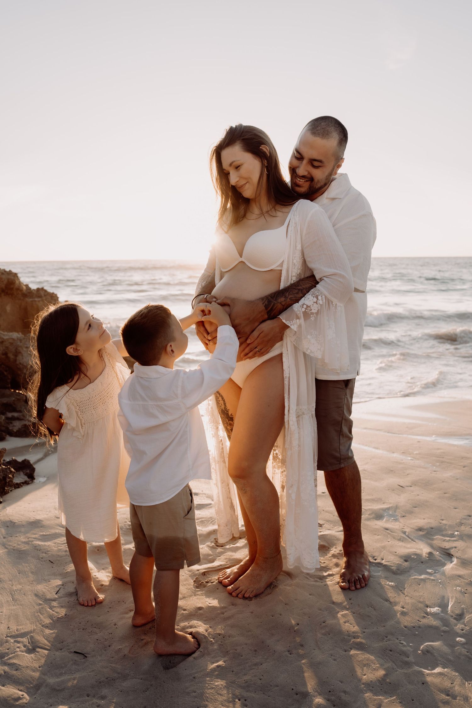 Pregnant woman in white bikini with husband and two children enjoying a beach photoshoot at golden hour.