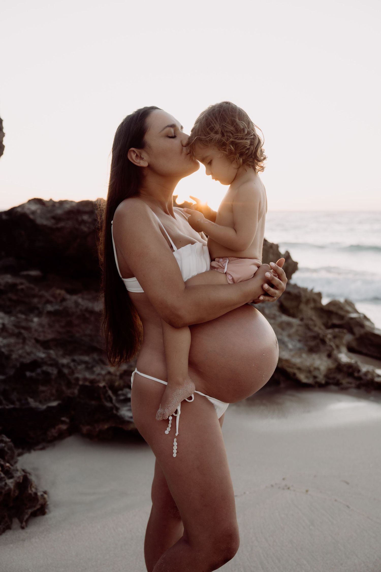 A pregnant woman in a white bikini holds a toddler while standing on a beach at sunset near rocky cliffs.