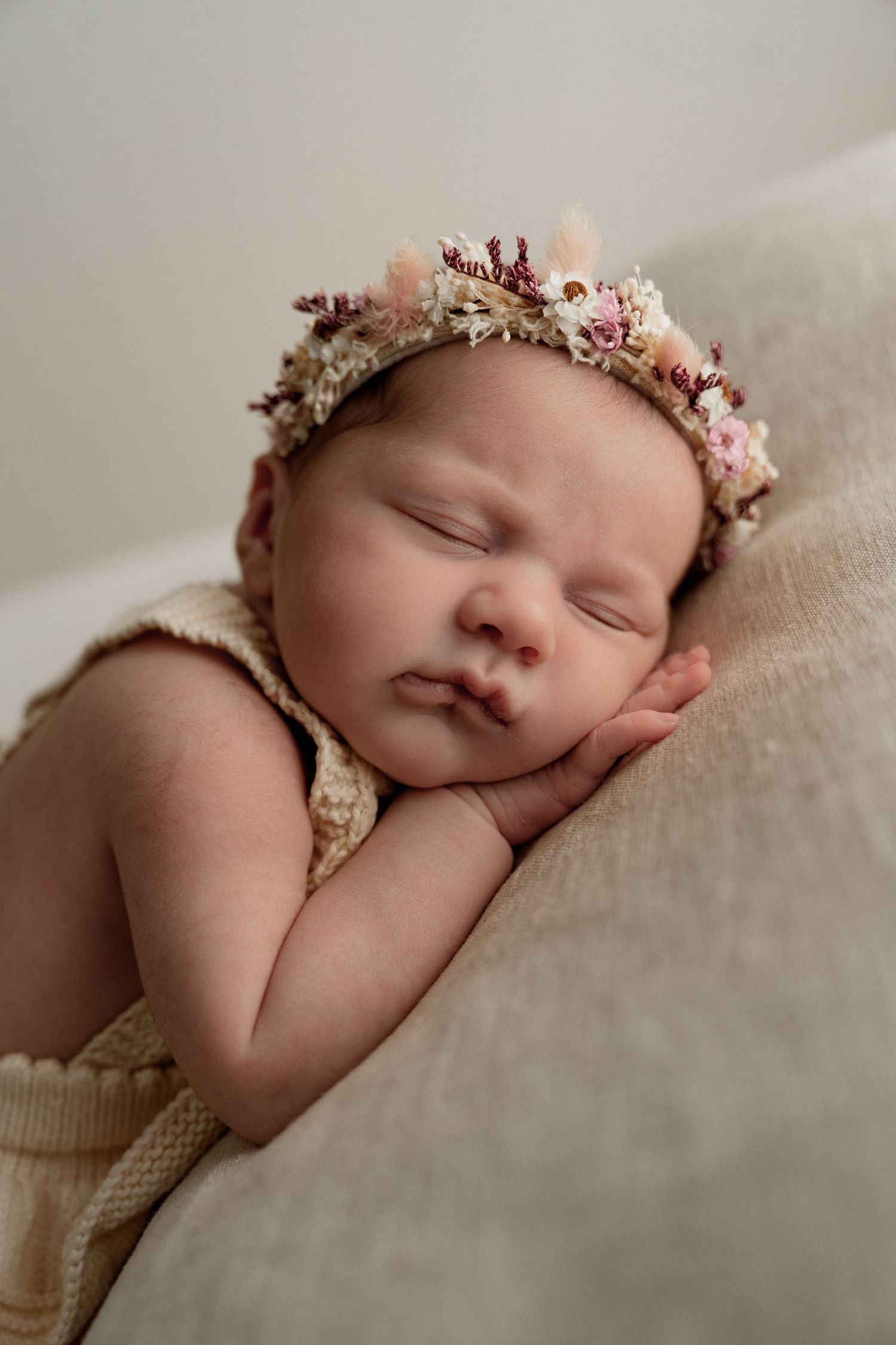 A peaceful newborn baby wearing a delicate floral crown lies sleeping on soft beige fabric.