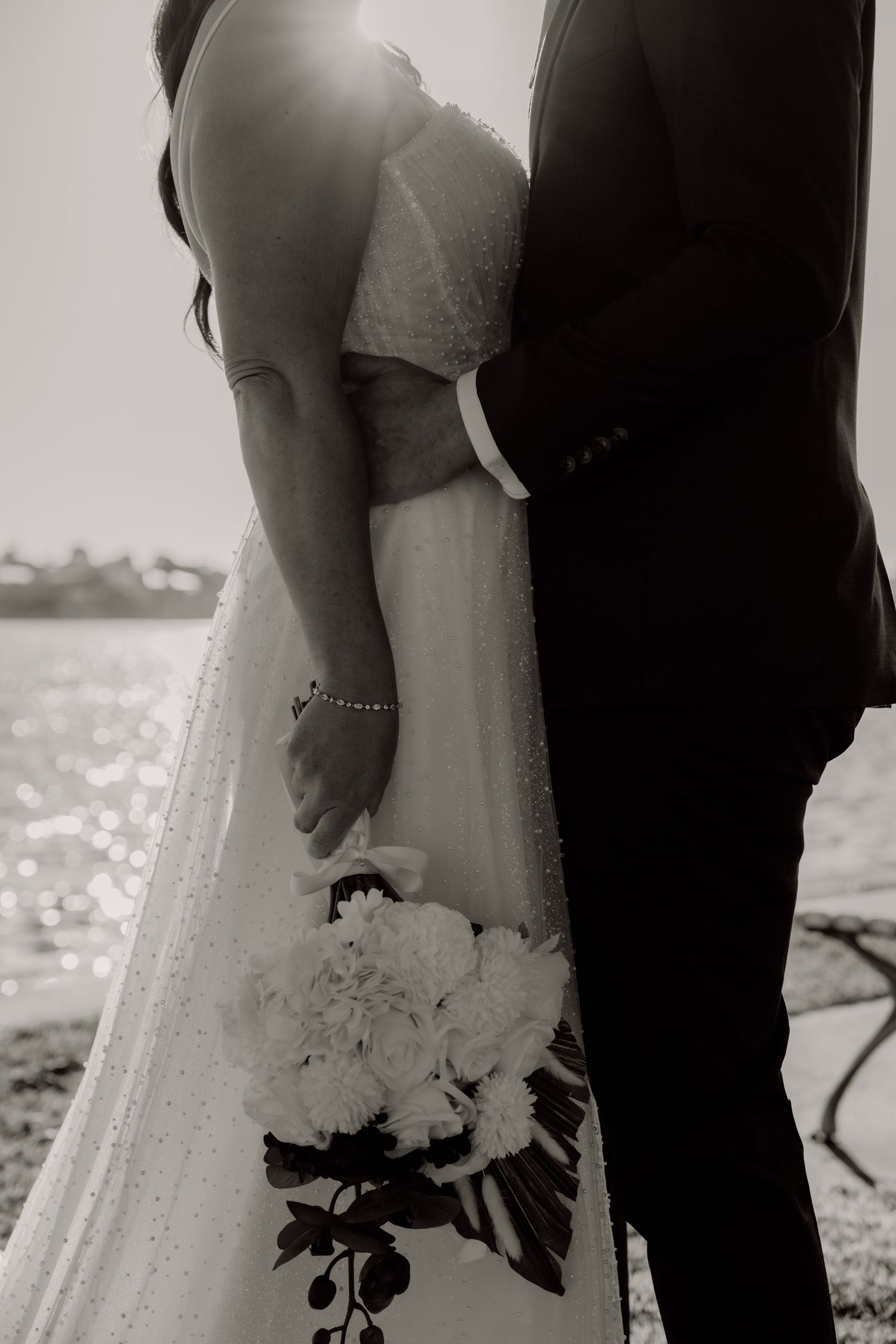 Black and white wedding photo showing bride in lace dress holding bouquet next to groom by waterfront.