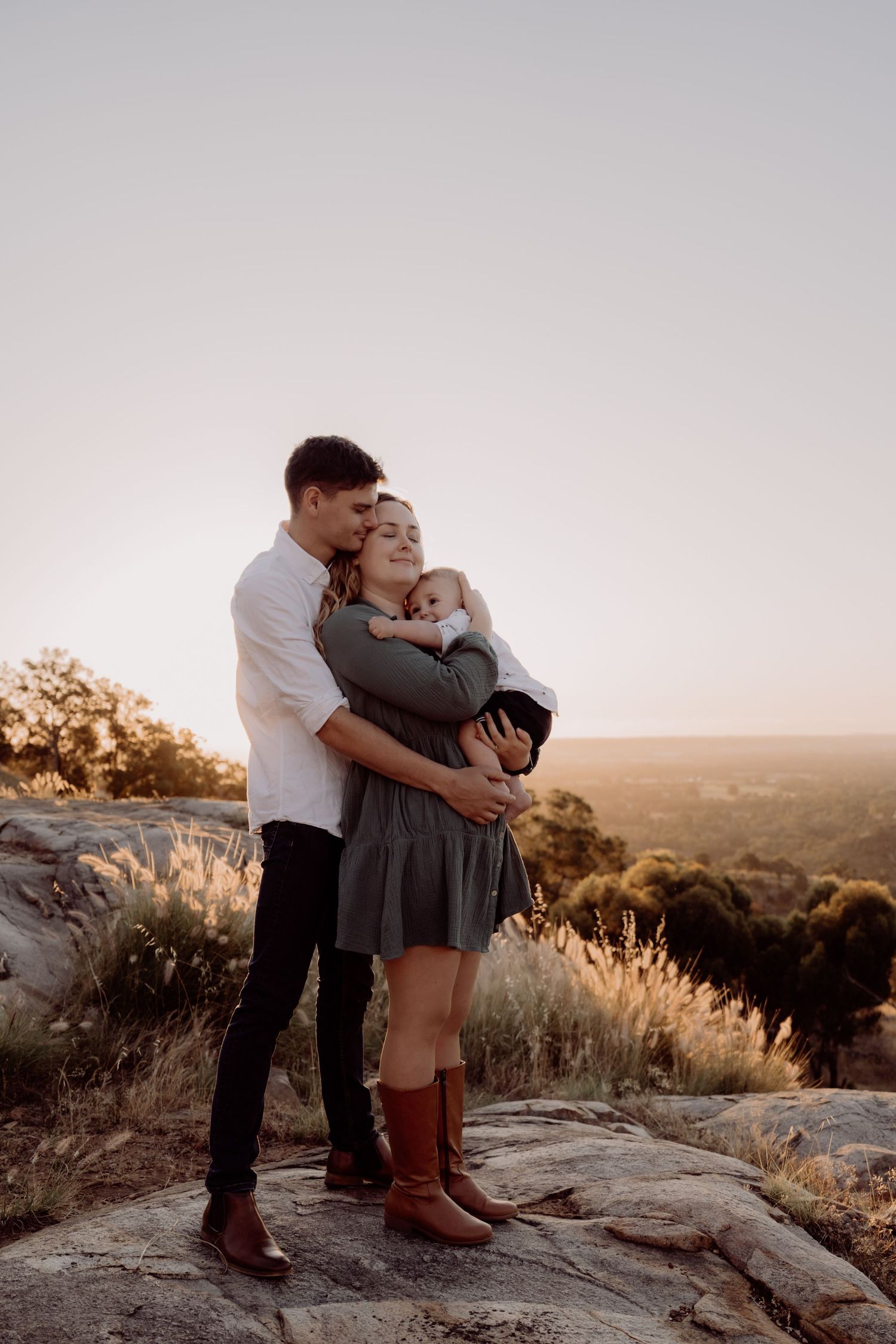A couple shares an intimate embrace while standing on rocks during sunset in a scenic mountain landscape.