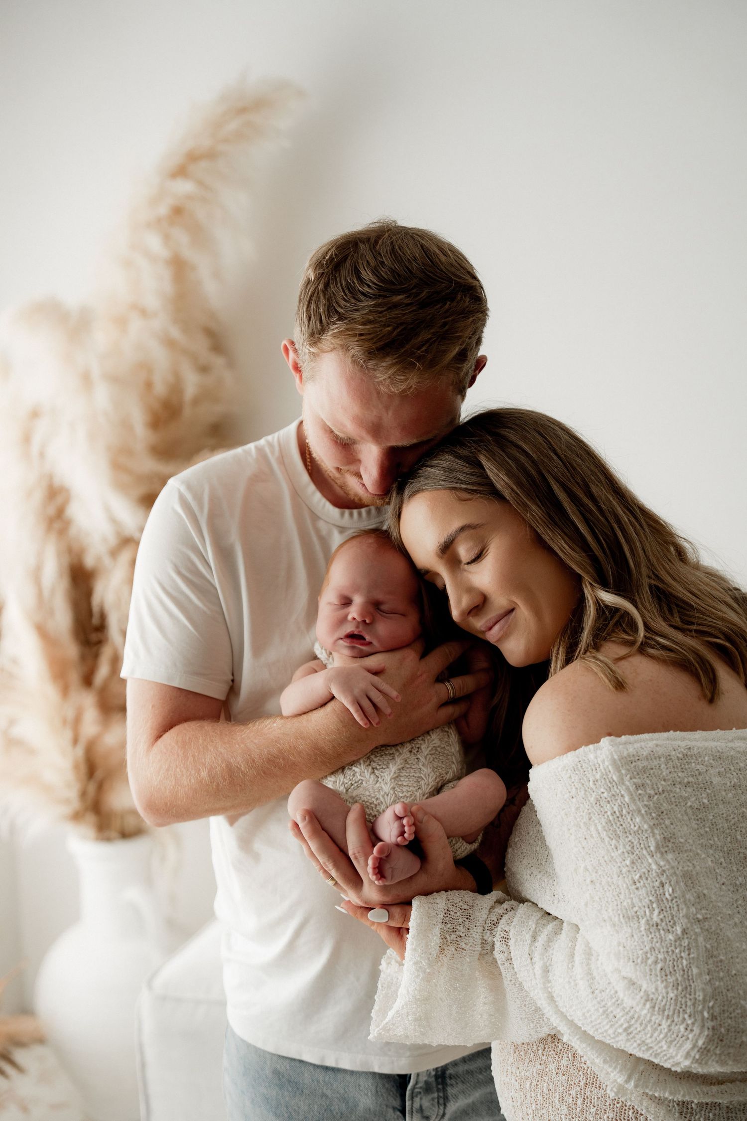 A couple tenderly holds their sleeping newborn baby wrapped in white, posing for a studio portrait.
