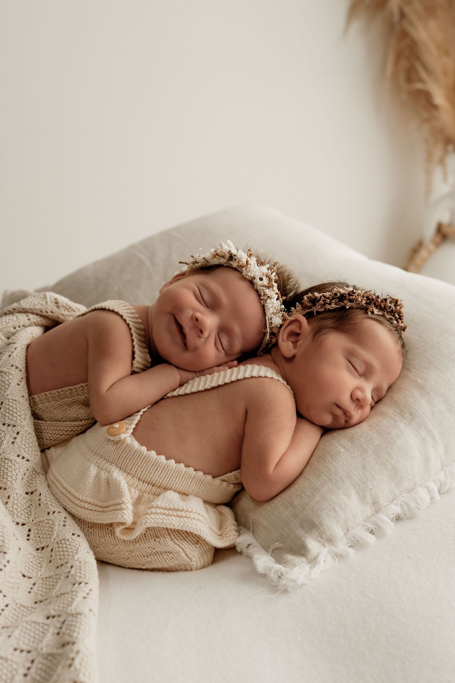 Sleeping newborn in cream knit wrap and floral crown photographed in soft natural light on white bedding.