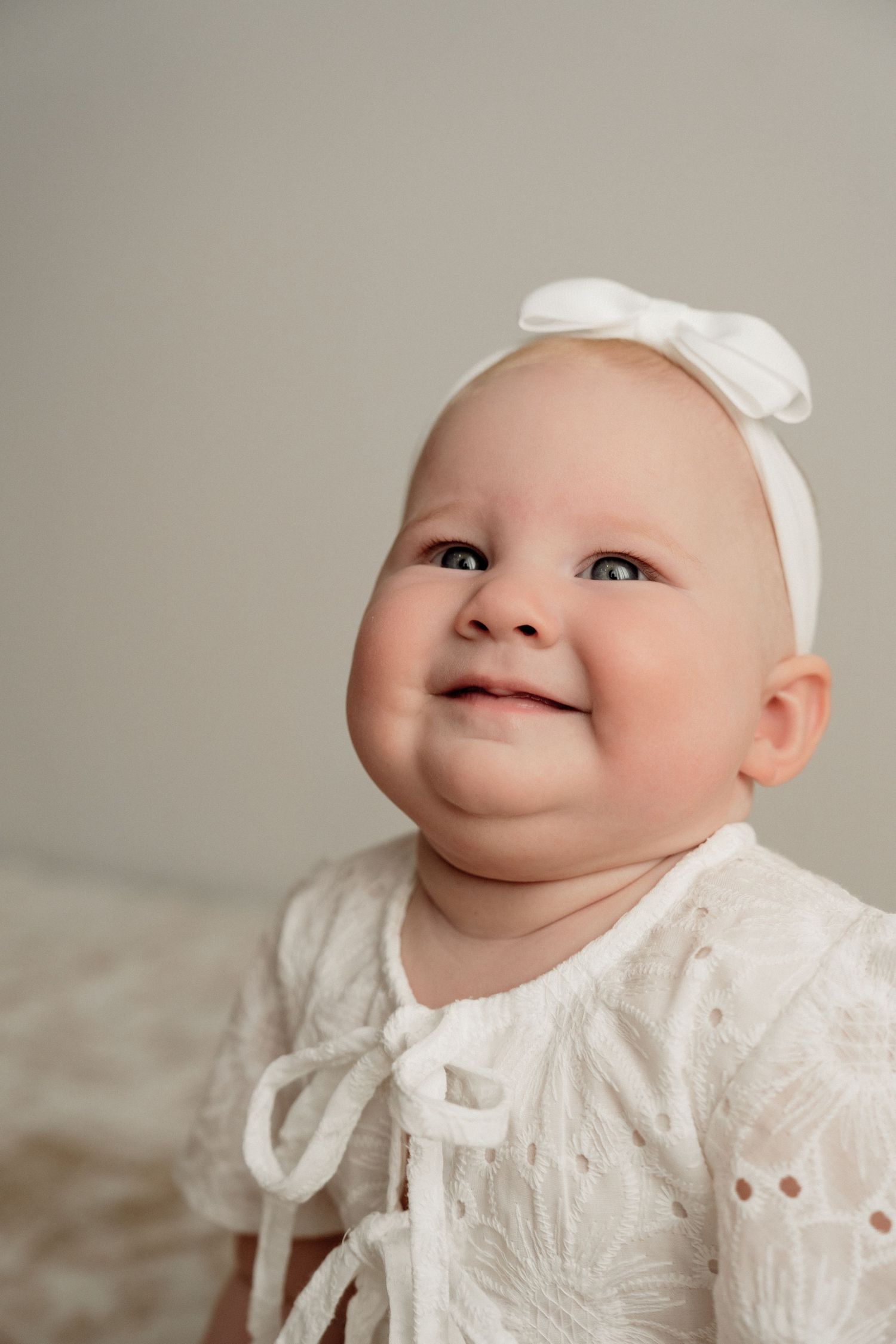 Baby wearing white lace dress smiles in series of closeup profile portraits against neutral background.