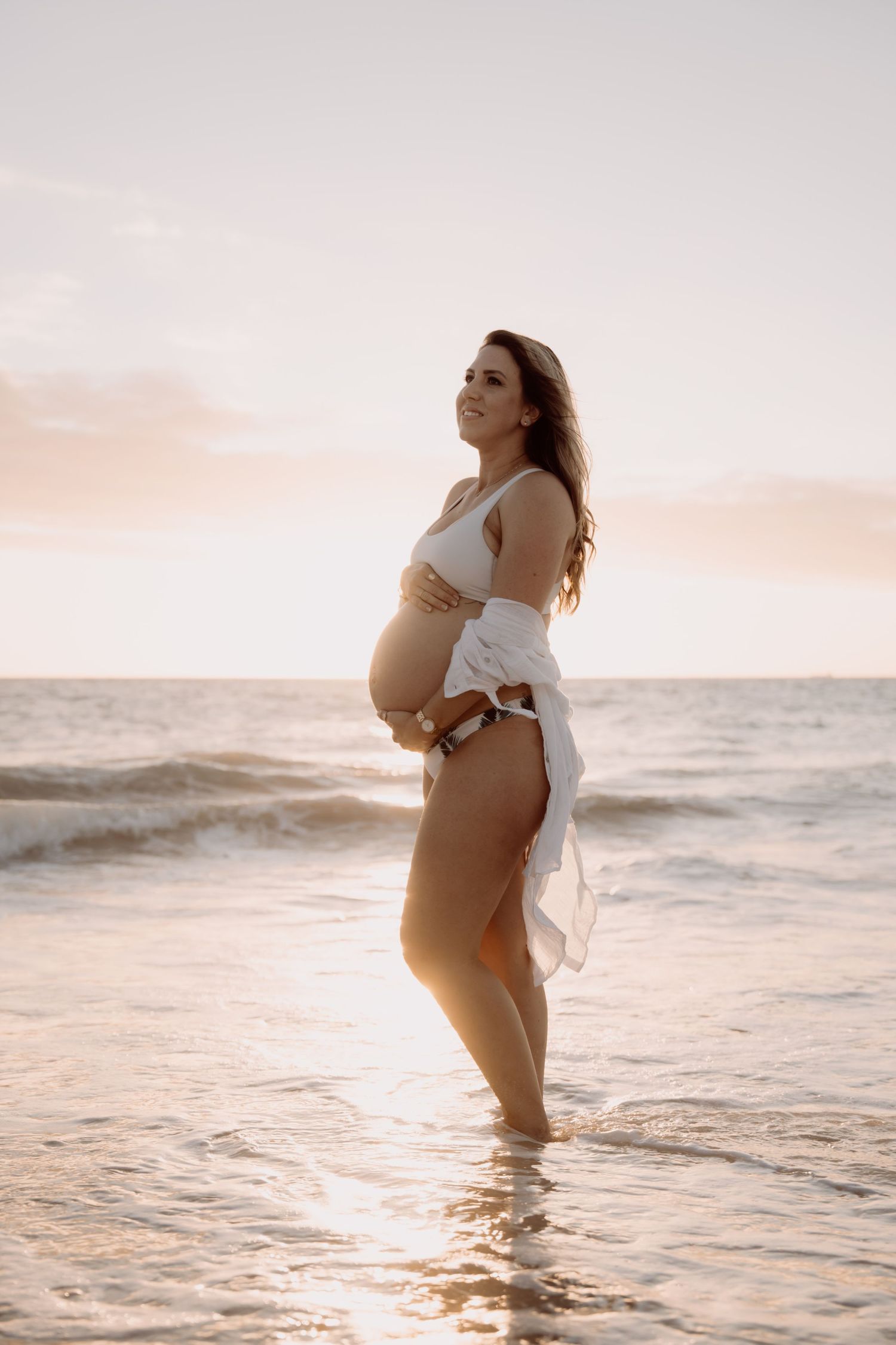Pregnant woman in white bikini and open shirt cradling her baby bump on a beach at golden sunset.