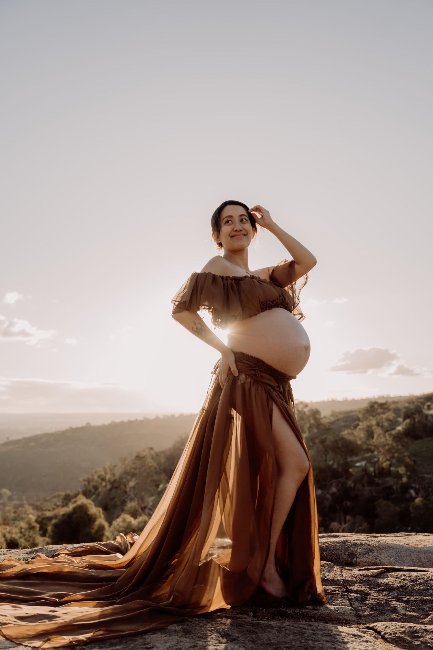 Golden hour maternity photoshoot on a rocky mountain overlook with flowing dress and dramatic sunset lighting.