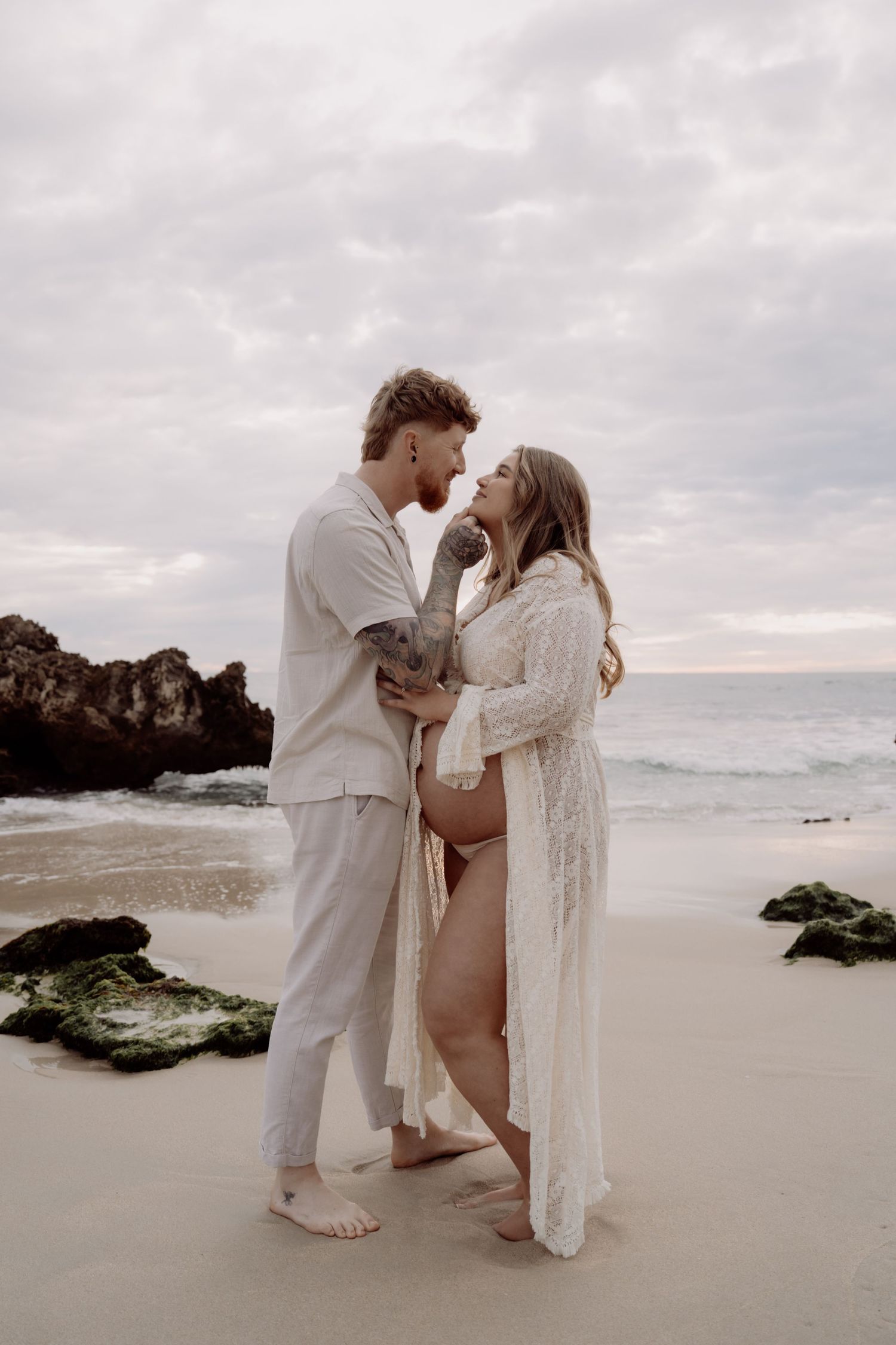 A couple shares an intimate moment on a misty beach at sunset, surrounded by rocky coastal scenery.