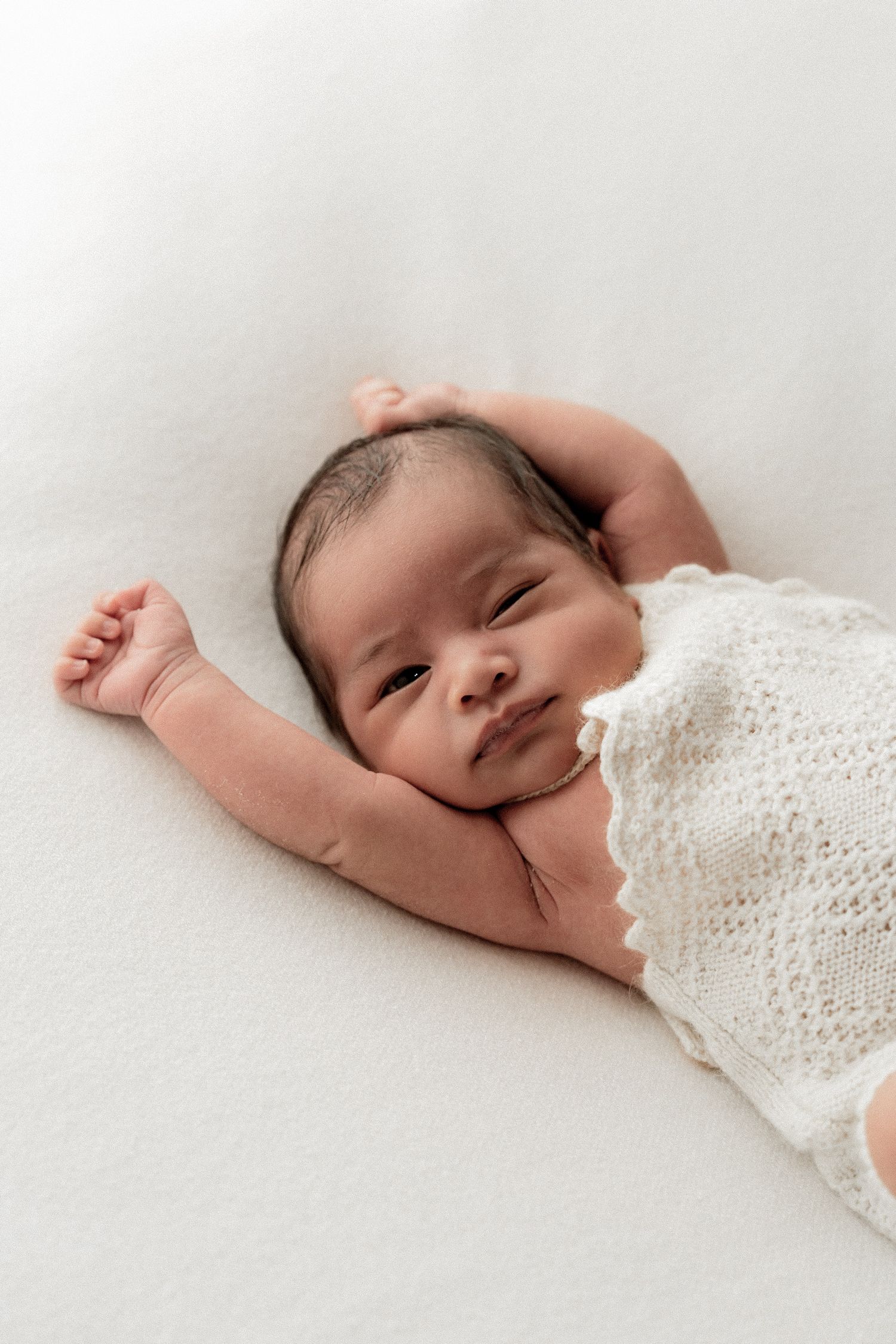 A sleeping newborn baby lies peacefully under a white knit blanket in a minimalist photography setup.