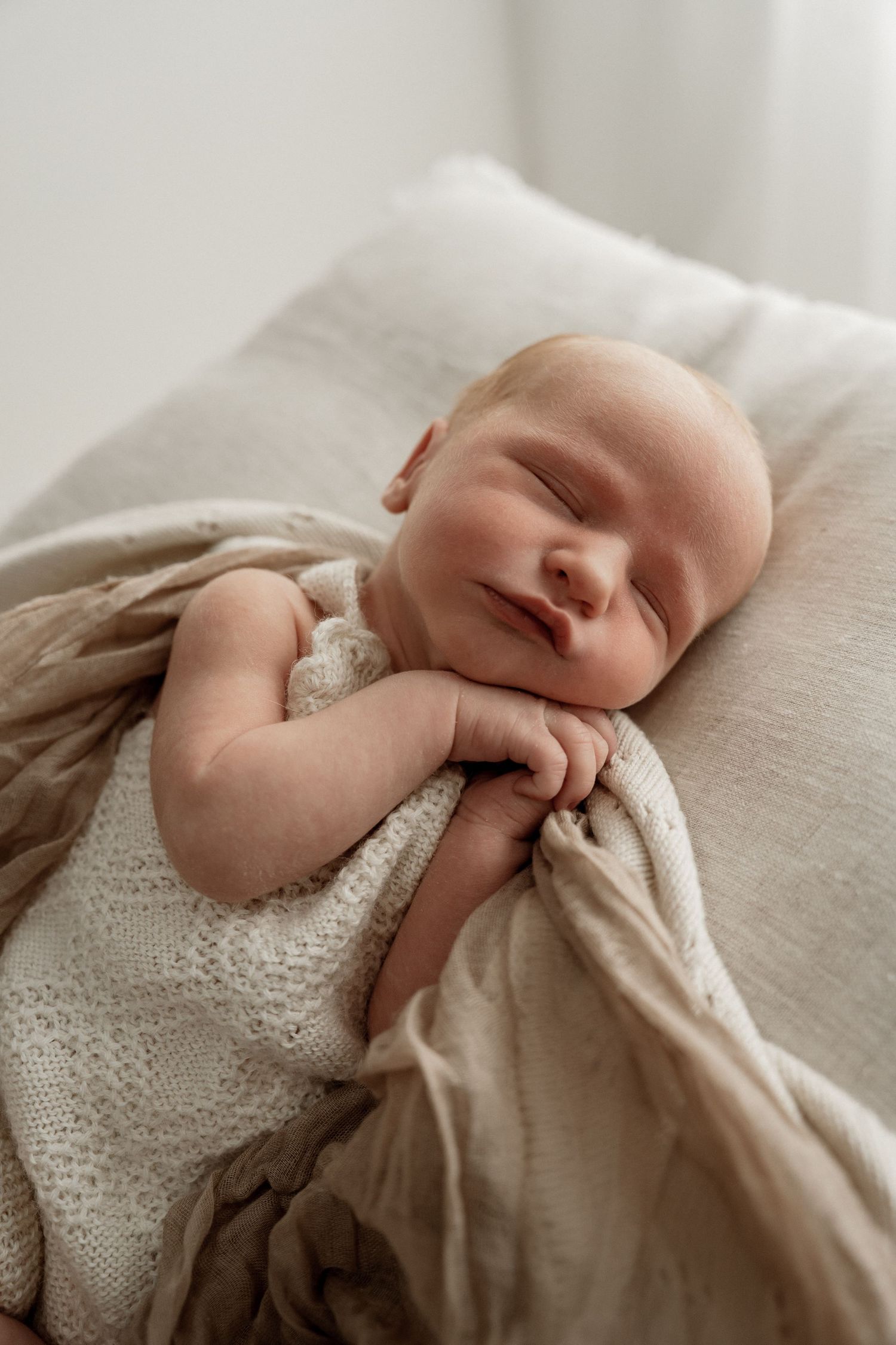 A sleeping newborn wrapped in soft beige fabric and lace rests peacefully in natural window light.