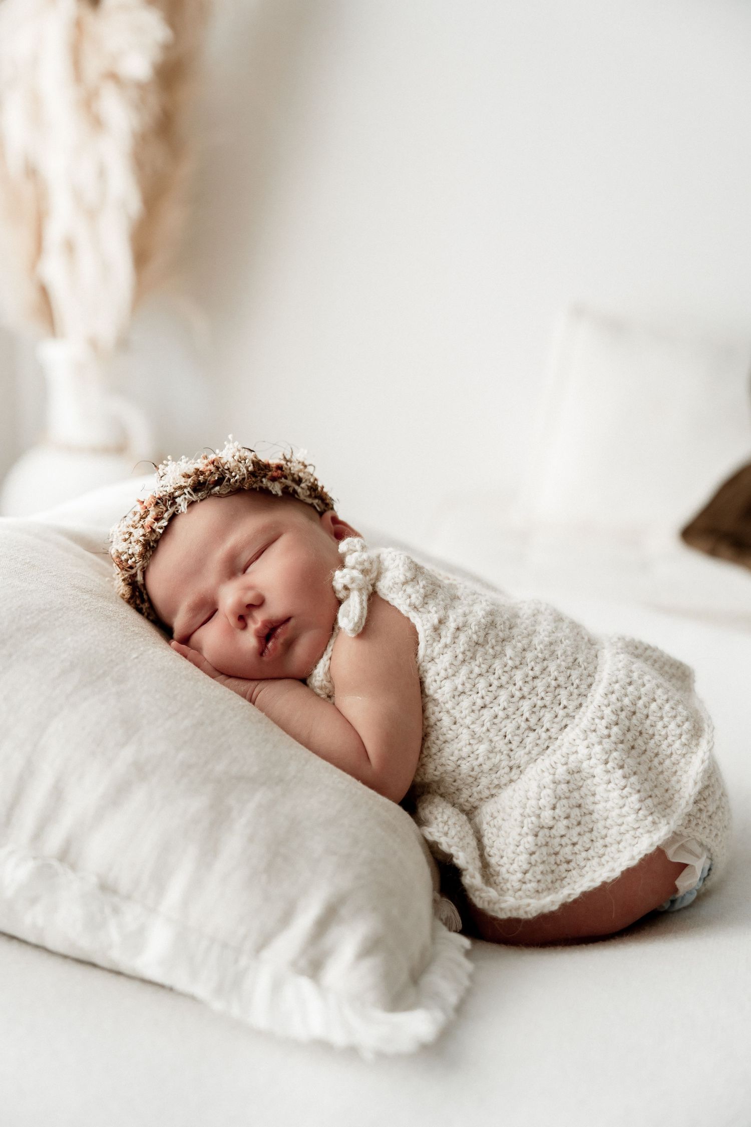 Sleeping newborn baby wearing a floral crown, wrapped in a cream knit blanket, resting on a linen pillow.
