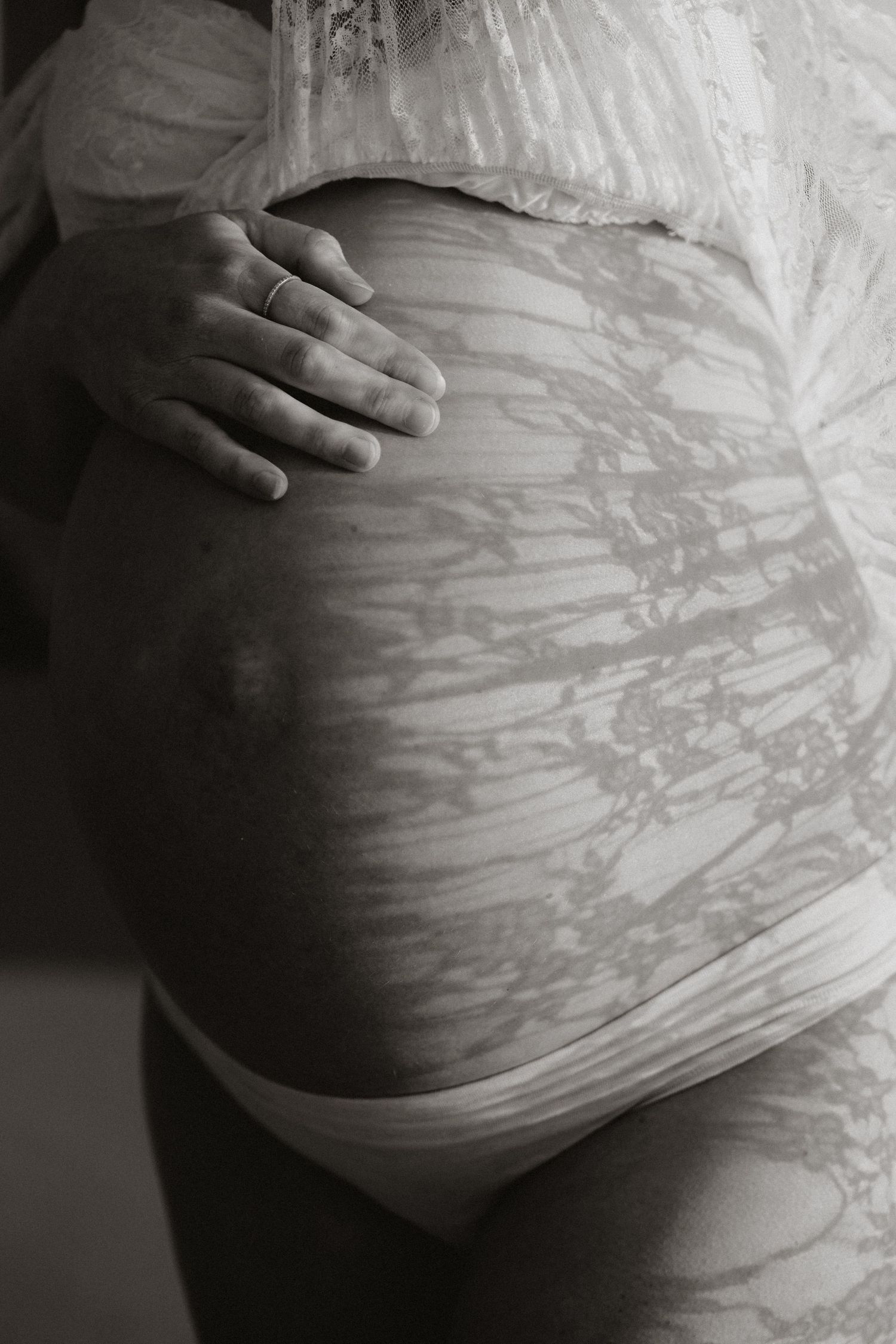 Black and white close-up of a pregnant woman's belly with lace fabric and hand resting gently on bump.