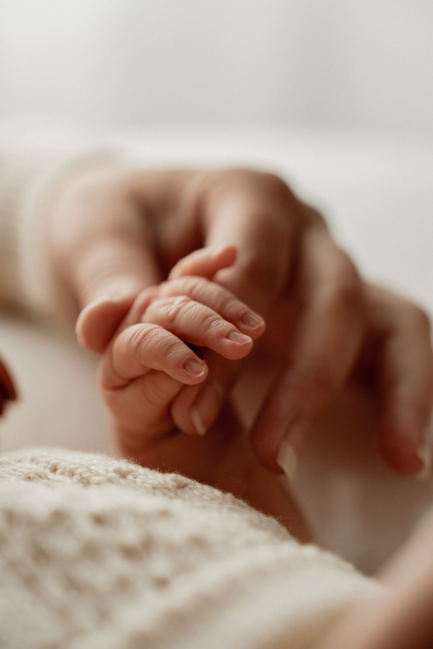 Tiny baby hand gently grasping adult finger against soft white fabric in warm natural lighting.