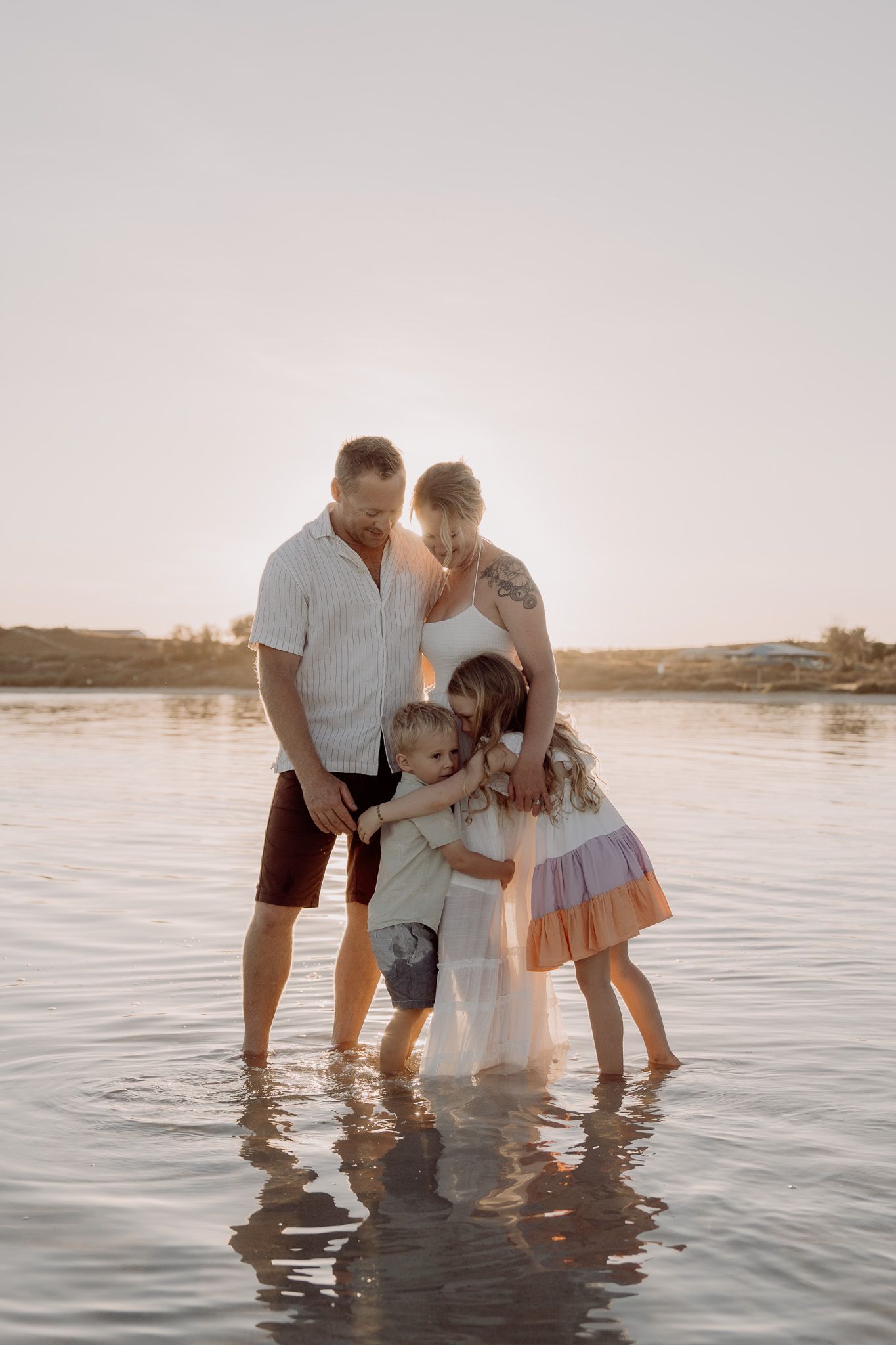 Family of four standing together in shallow water at beach during sunset.