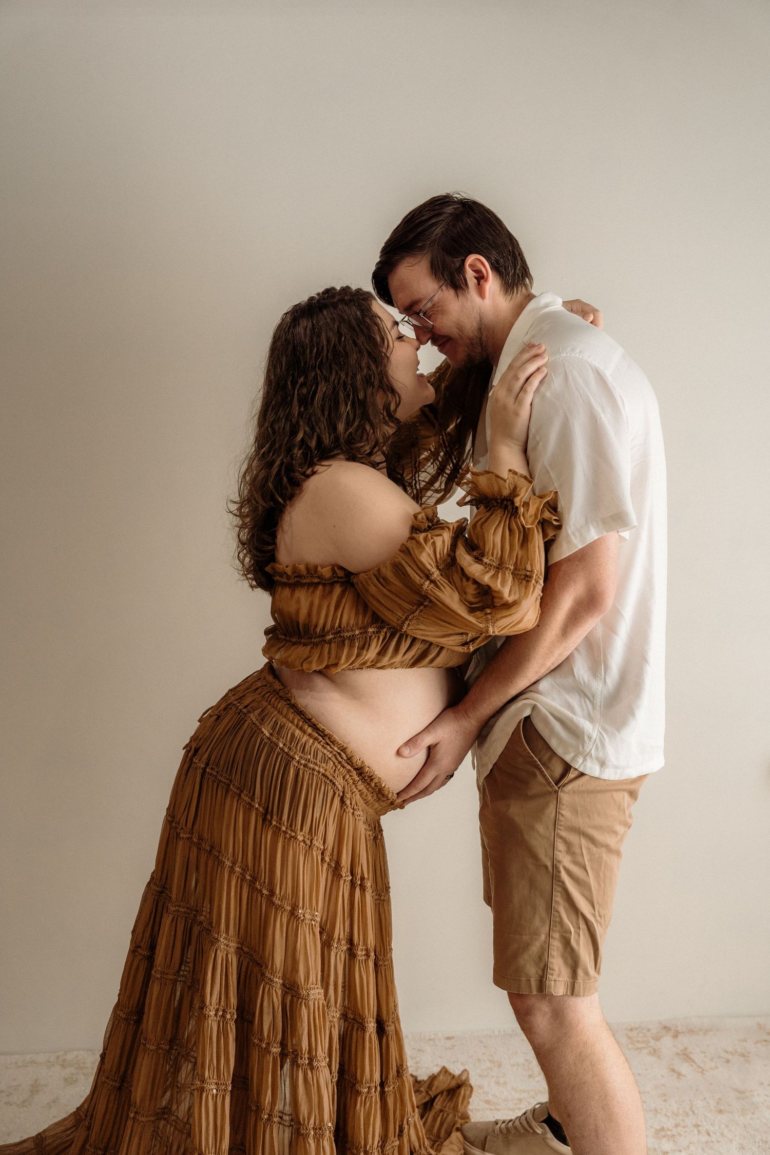 Expectant couple in intimate maternity photoshoot with woman wearing flowing brown tiered dress against neutral backdrop.