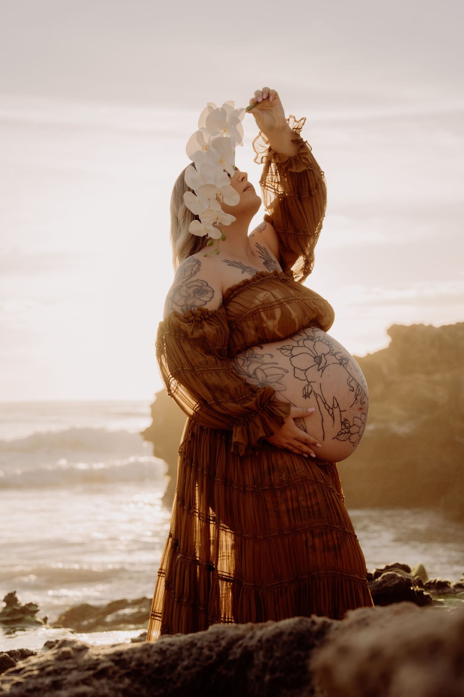 Silhouette of a pregnant woman in a flowing dress standing on a beach at sunset, holding paper flowers aloft.