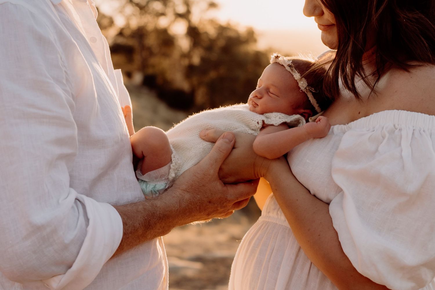 A newborn baby sleeps peacefully between its parents during a golden hour outdoor photo session.