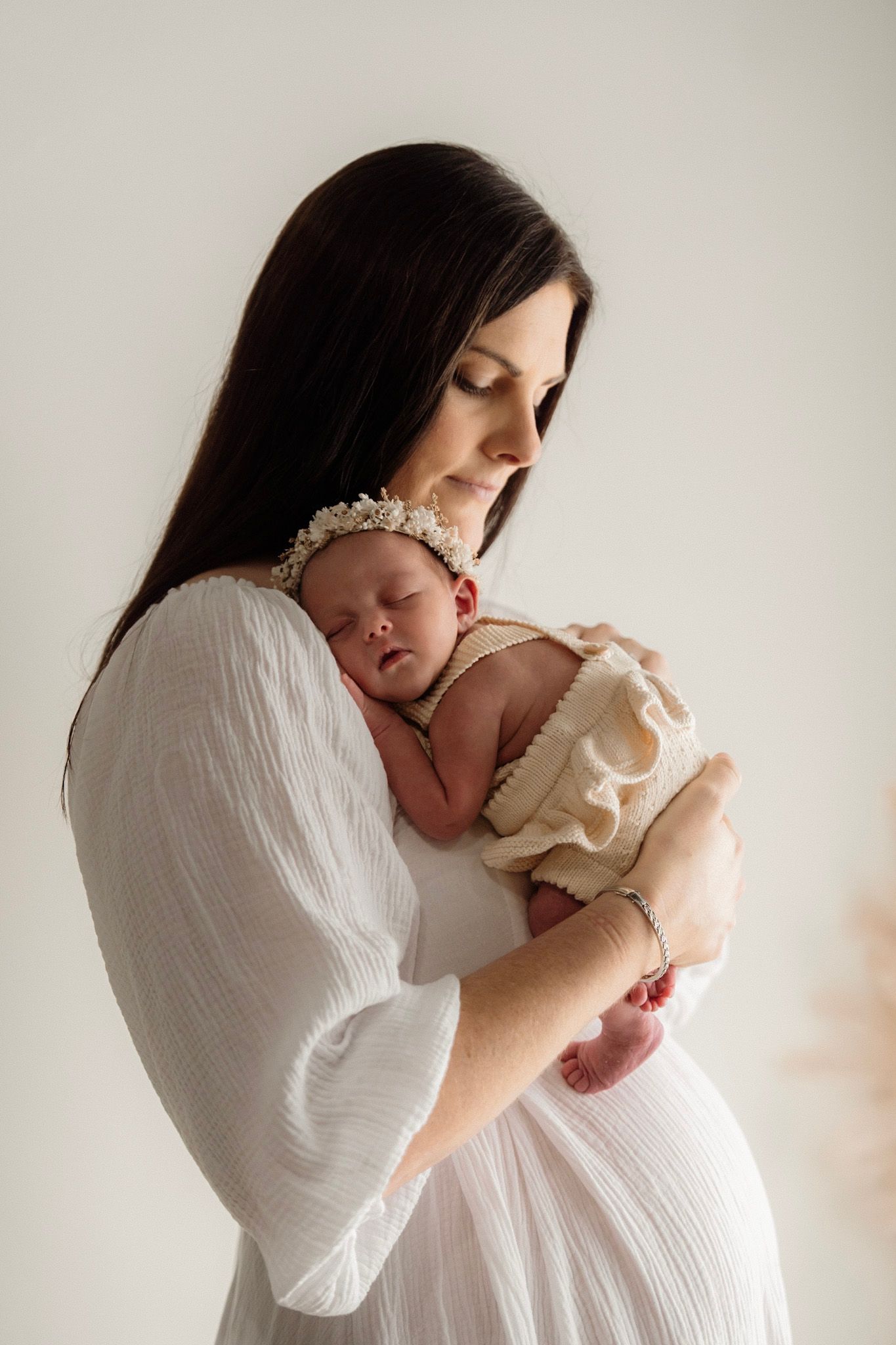 A mother wearing a white gauzy blouse holds and kisses her newborn baby in a minimalist light-filled setting.