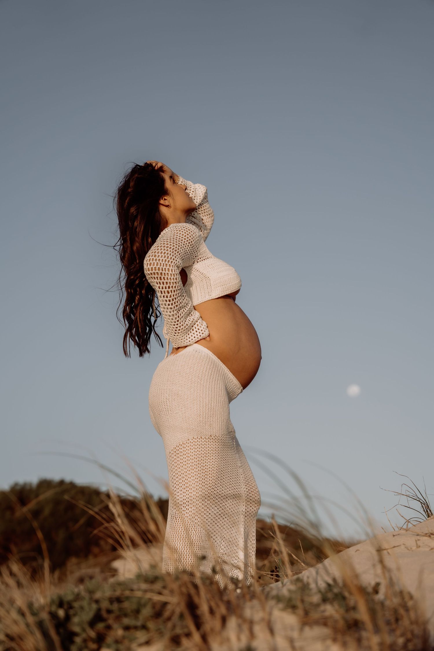 Pregnant person in flowing white crochet dress poses on beach dunes at sunset.