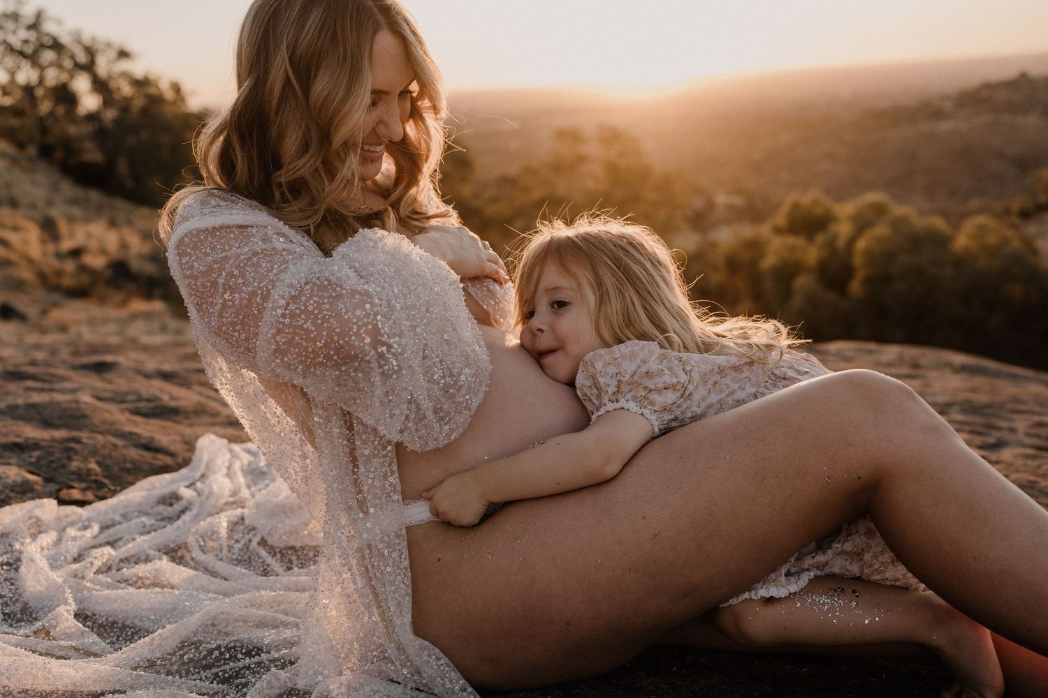 A tender maternity moment captured at sunset as parent and child embrace on a sandy hill in white lace clothing.