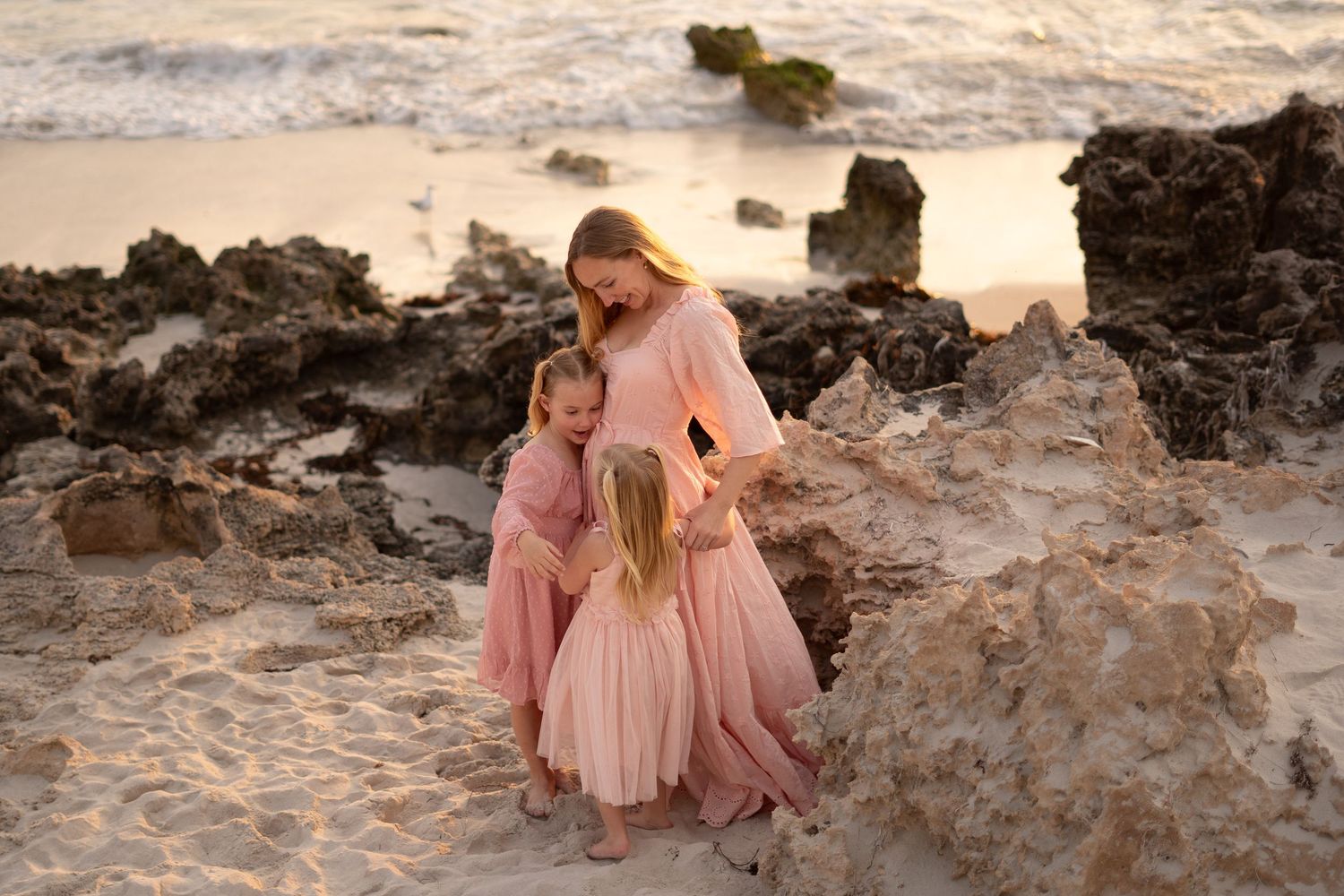 Three people in matching pink dresses stand among rocky coastal formations during a golden sunset.