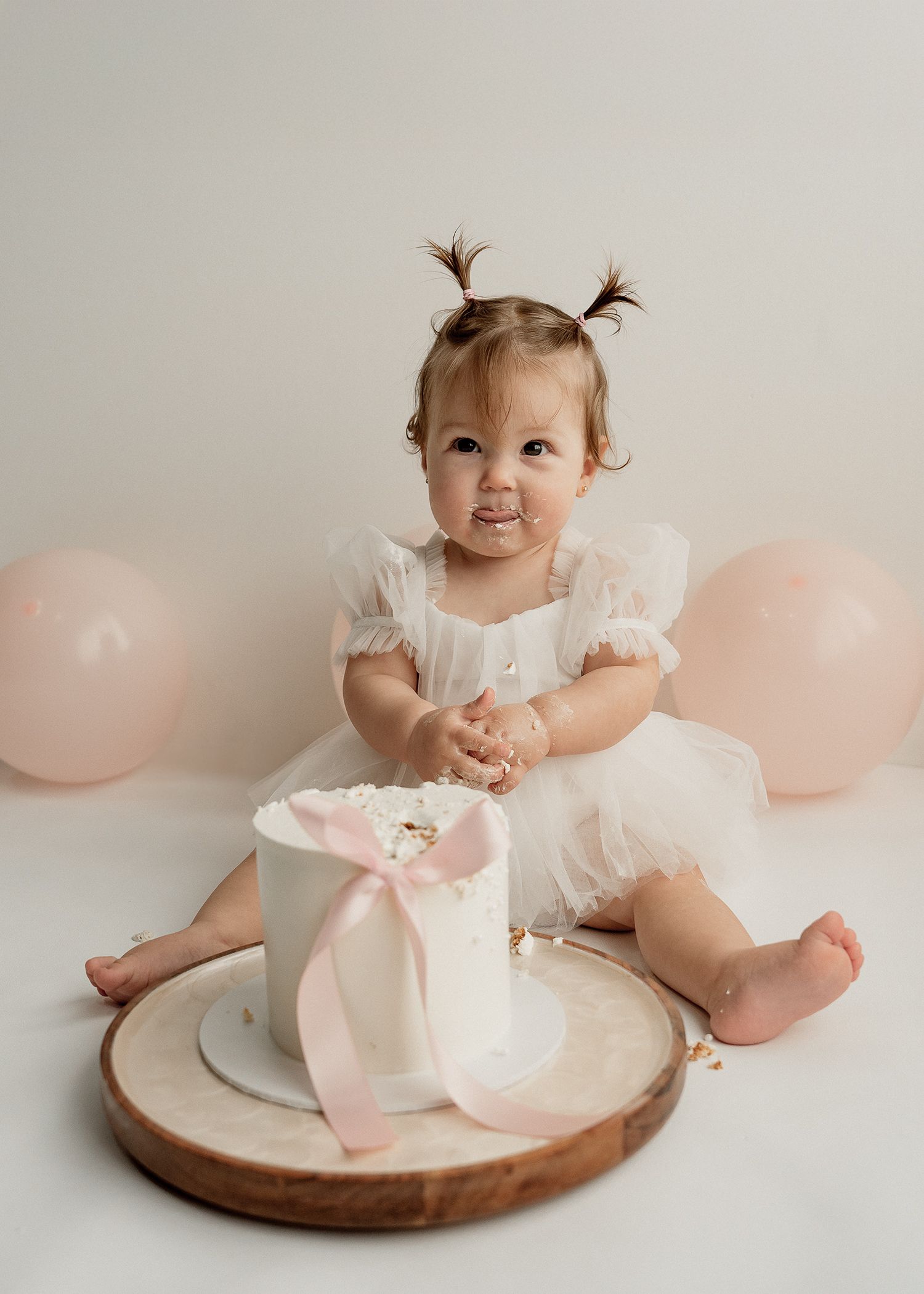 Baby in white dress with pigtails sitting on wooden platter with pink balloons in background for first birthday photo shoot.