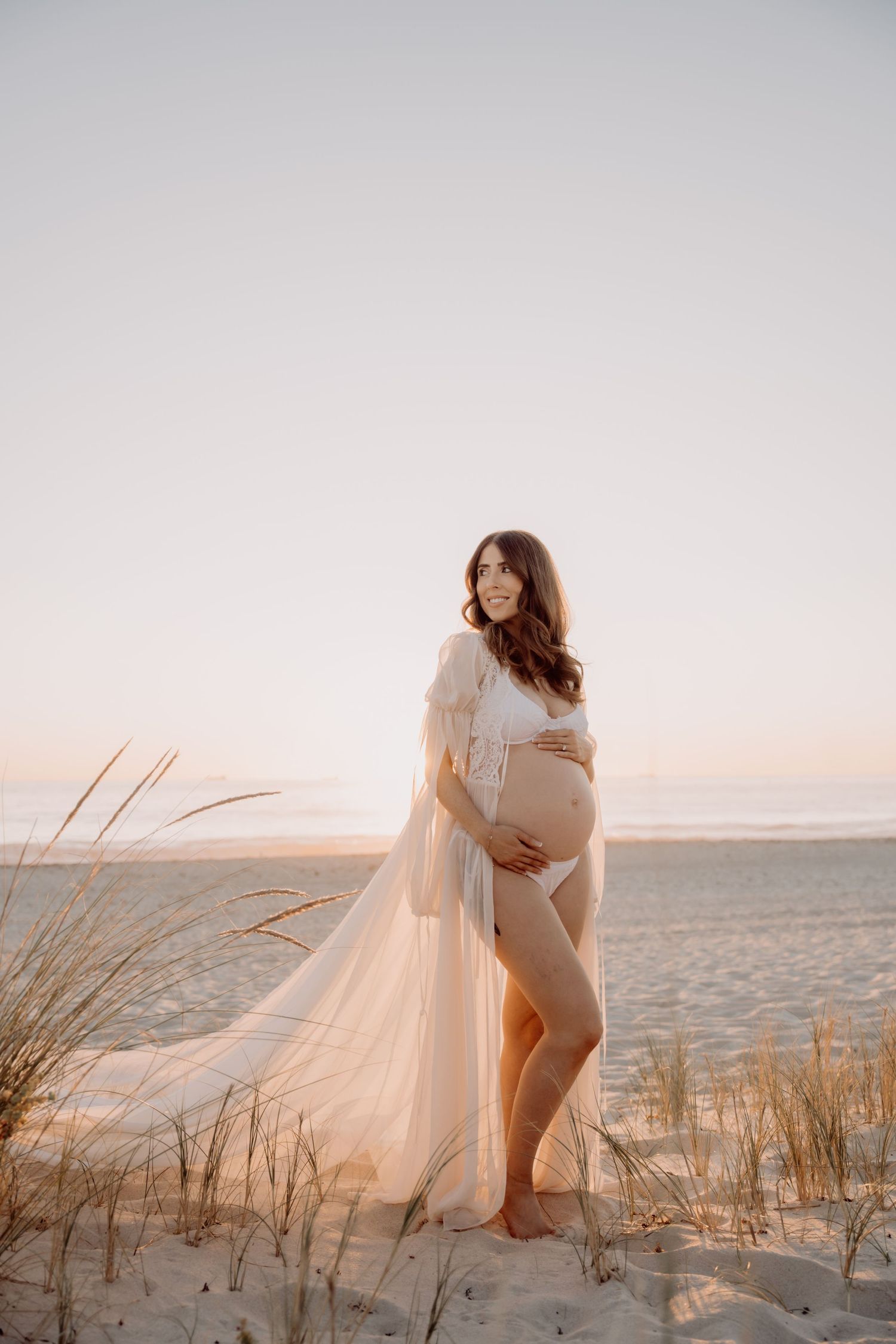 Pregnant woman in flowing white sheer gown poses on sandy beach at golden sunset, showcasing baby bump.