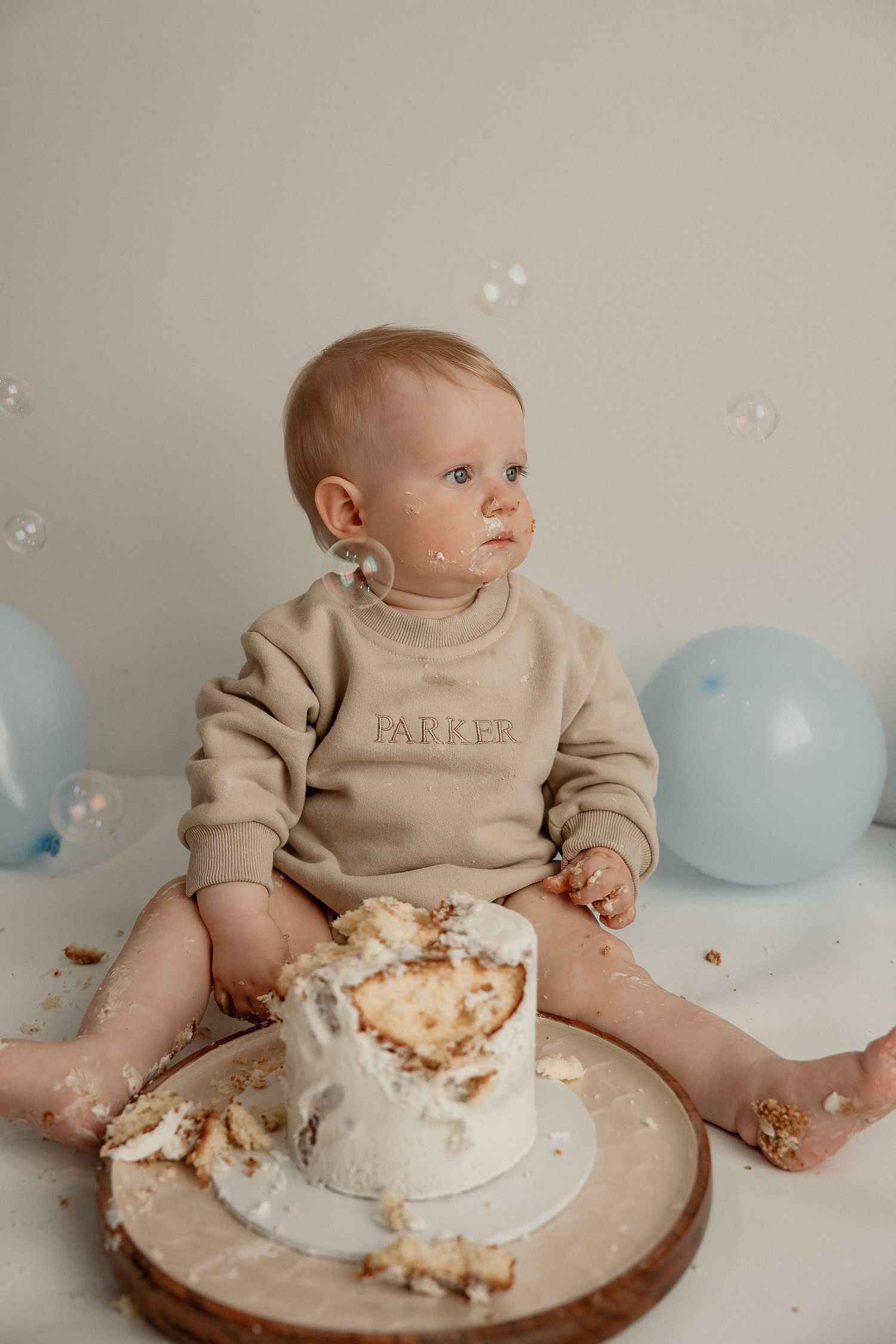 Baby in beige outfit sits beside smashed birthday cake with blue balloons in background during cake smash photoshoot.