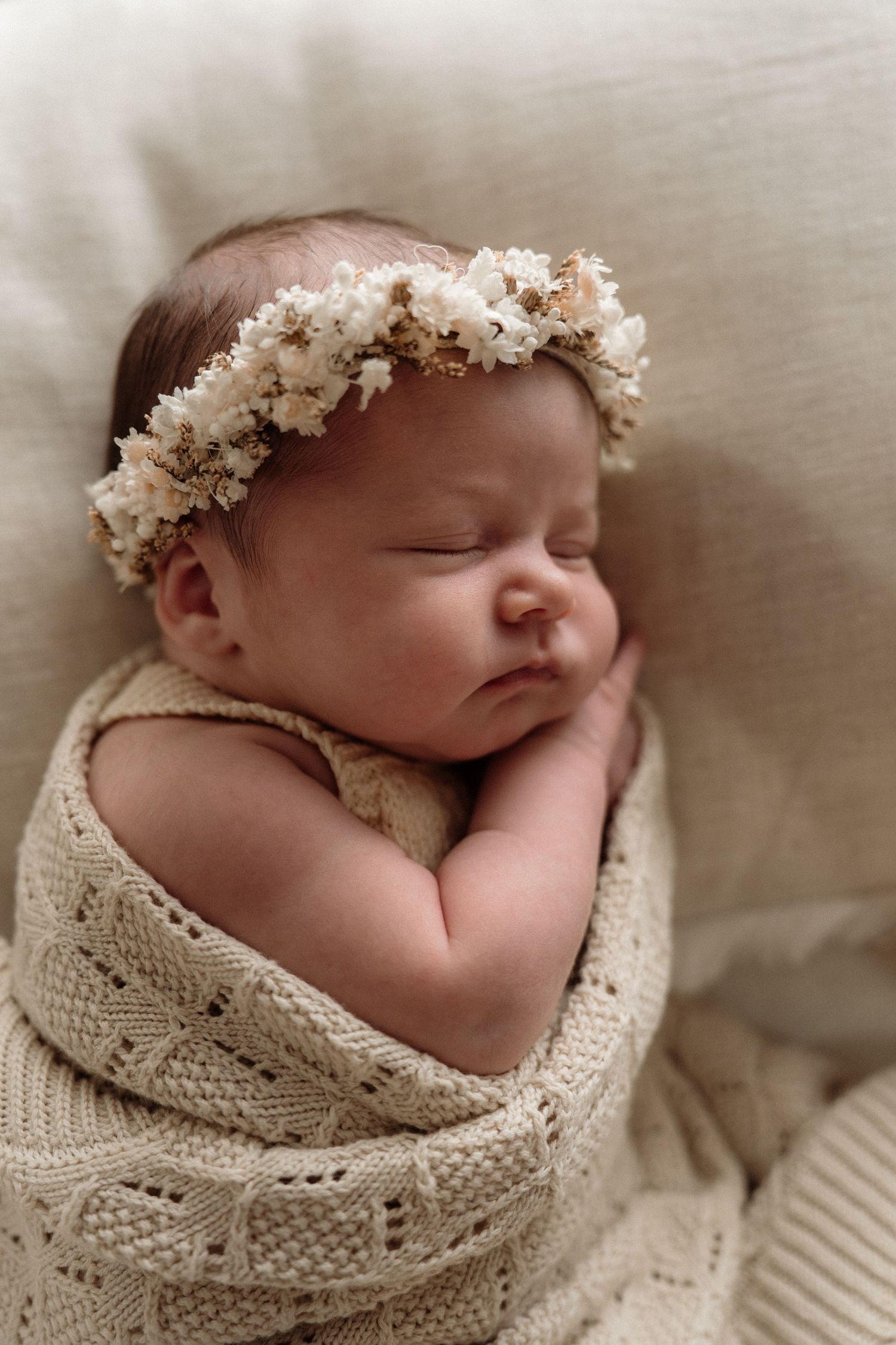 Peacefully sleeping newborn wrapped in cream knit blanket wearing delicate white floral crown headband.
