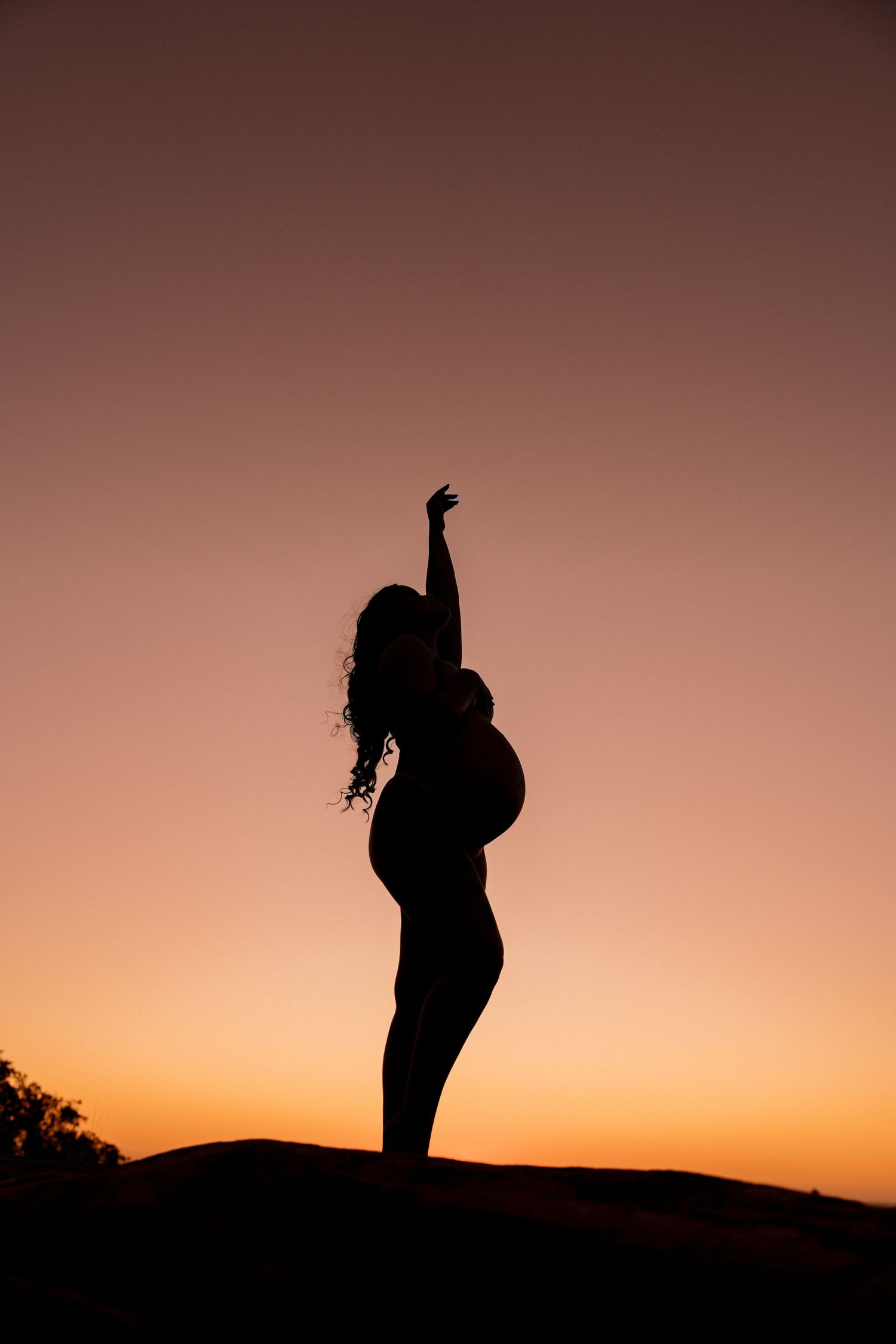 Silhouette of pregnant woman with curly hair striking a pose against vibrant orange sunset sky.