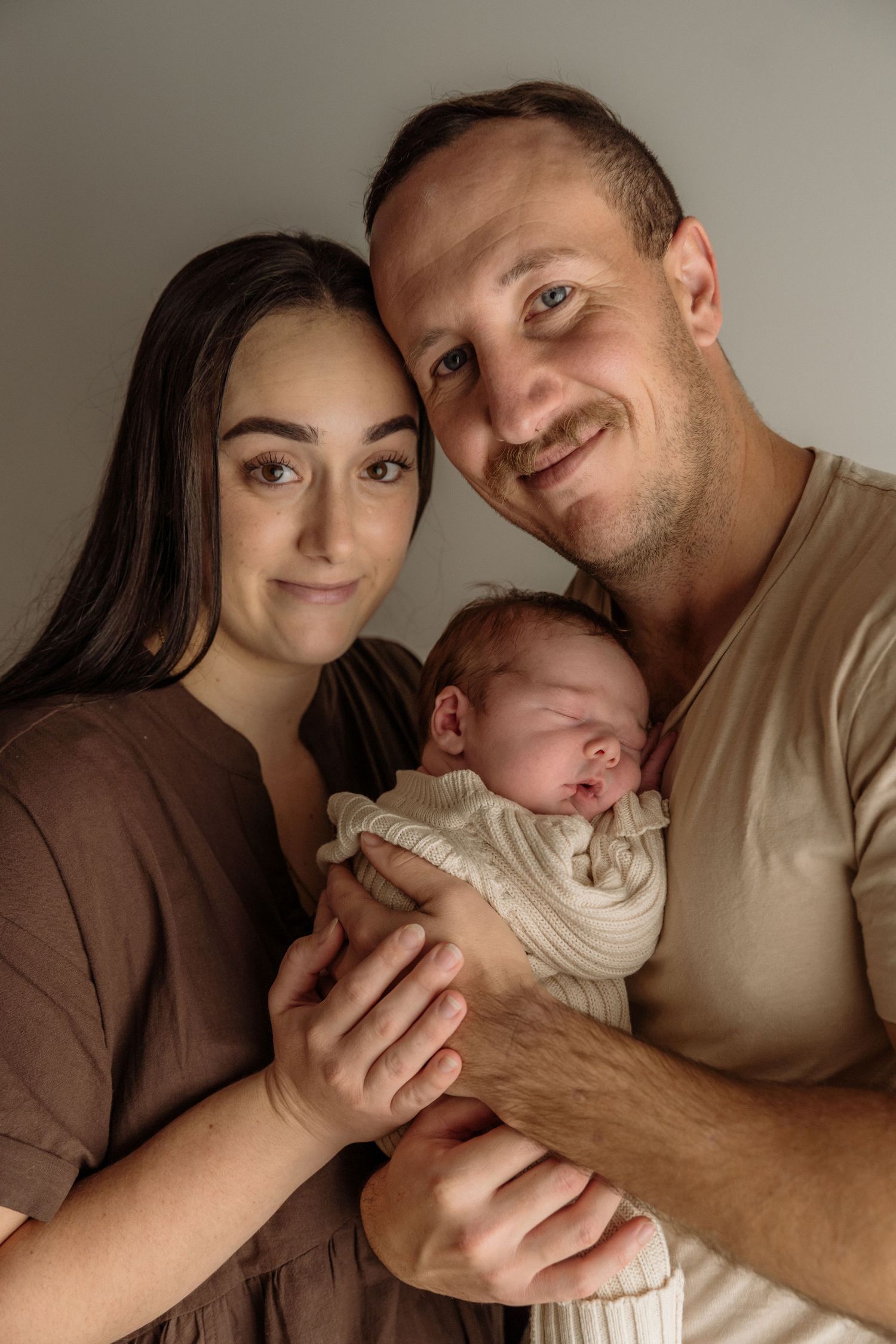 A couple holds their sleeping newborn baby wrapped in white cloth against a light neutral background in a tender moment.