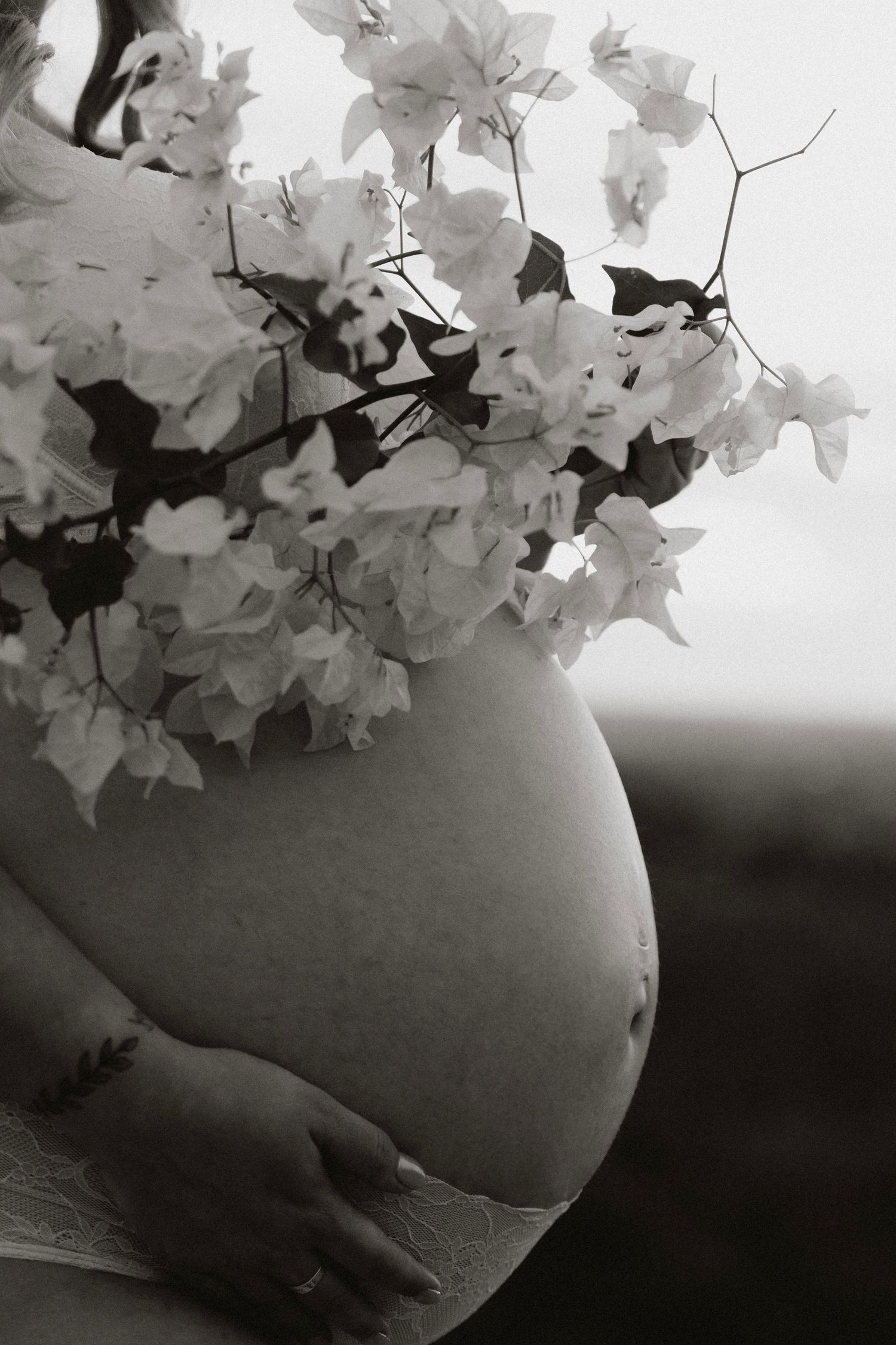 Black and white photo of a pregnant woman holding bougainvillea flowers over her bare baby bump.
