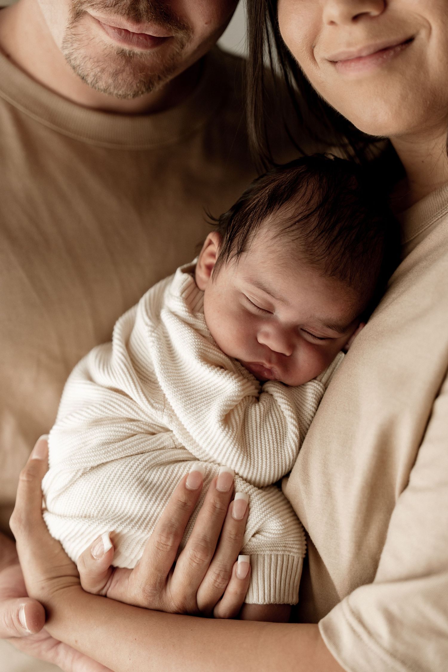 A sleeping newborn baby in a cream knit outfit nestles between two adults in beige clothing.