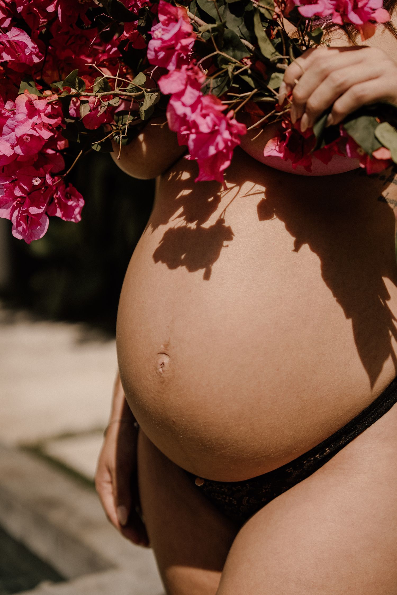 A pregnant woman's belly is adorned with pink bougainvillea flowers and shadows in warm natural lighting.