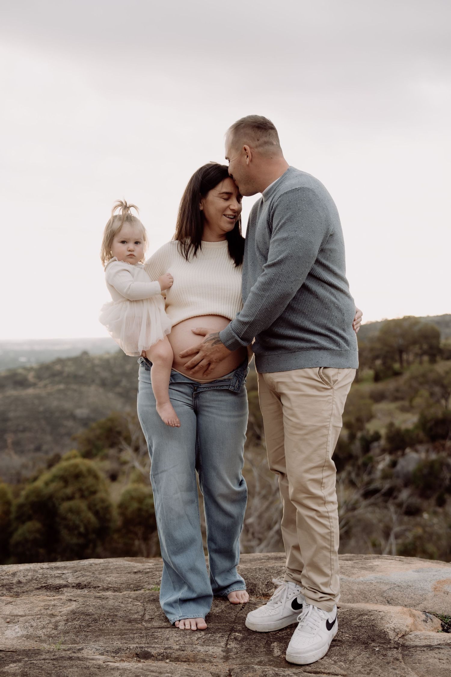 Pregnant woman standing on rocky overlook with husband kissing her head and toddler daughter in pink dress.