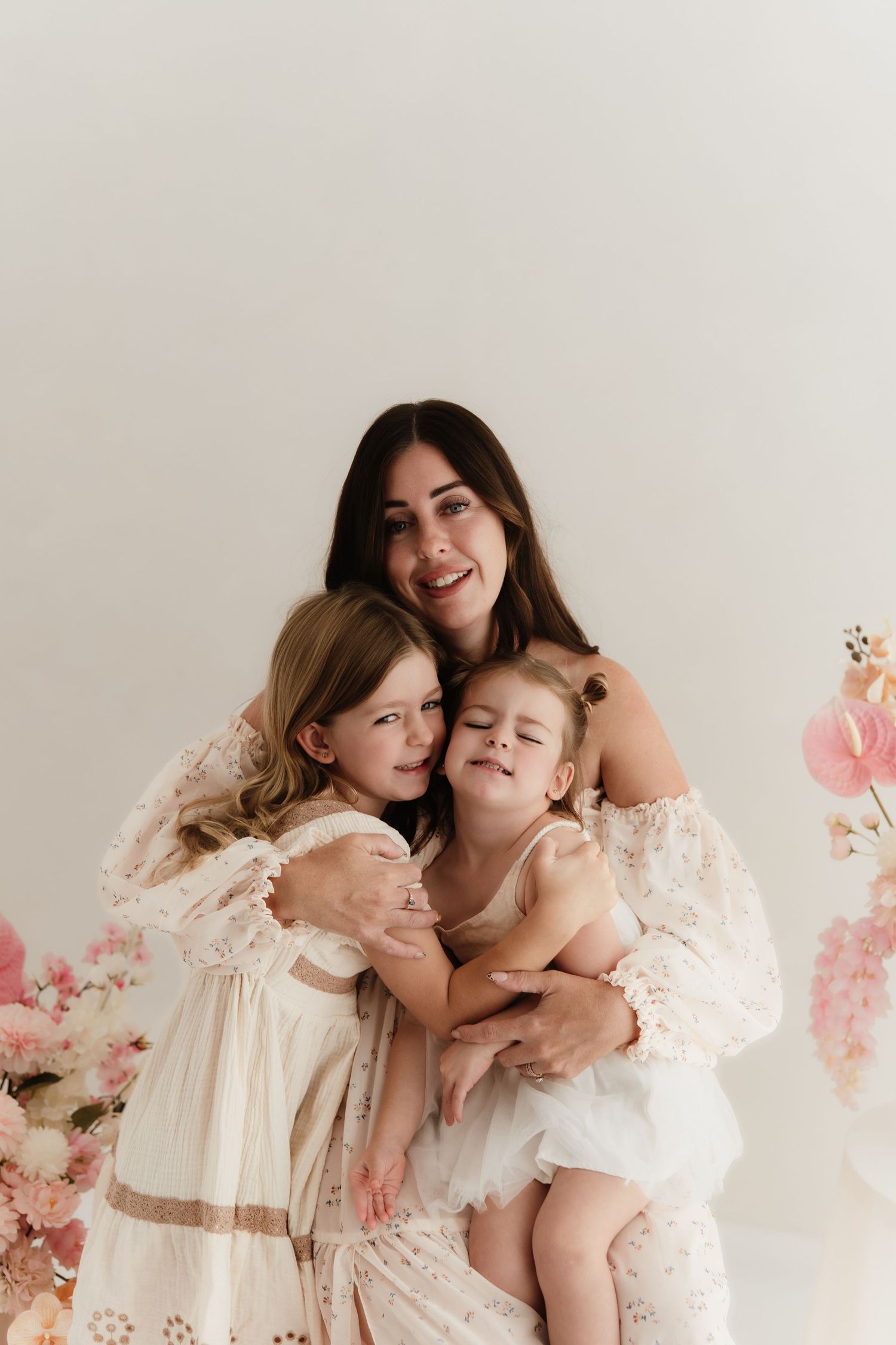 A smiling brunette mother in a floral dress hugs her two young daughters in white dresses against a soft pink floral backdrop.