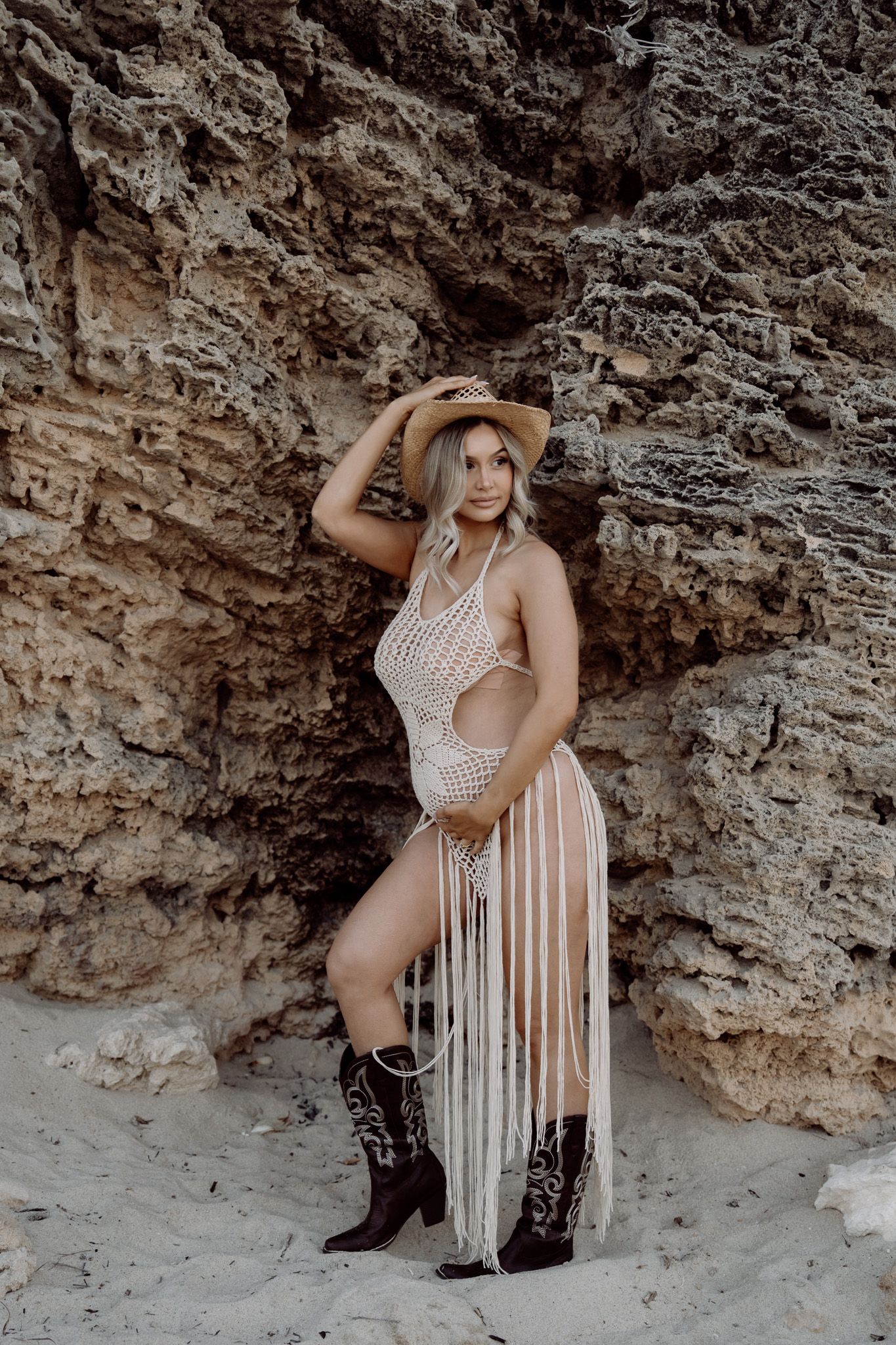 A model in a white fringe bodysuit and black boots poses against a rocky cave wall in a desert setting.