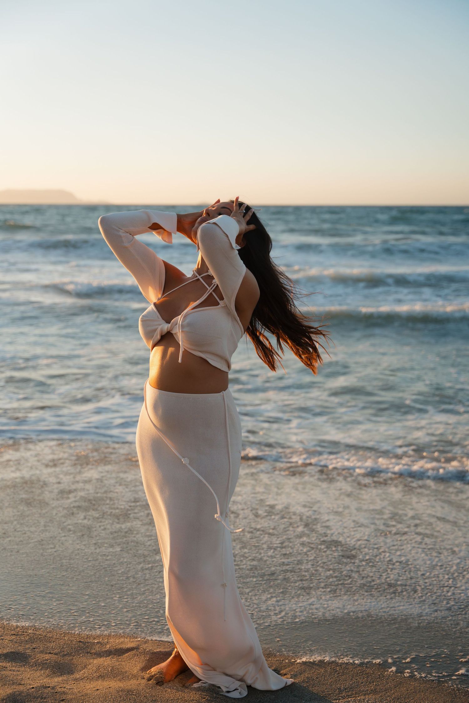 Woman in flowing white outfit dances on beach at sunset with ocean waves and golden light in background.