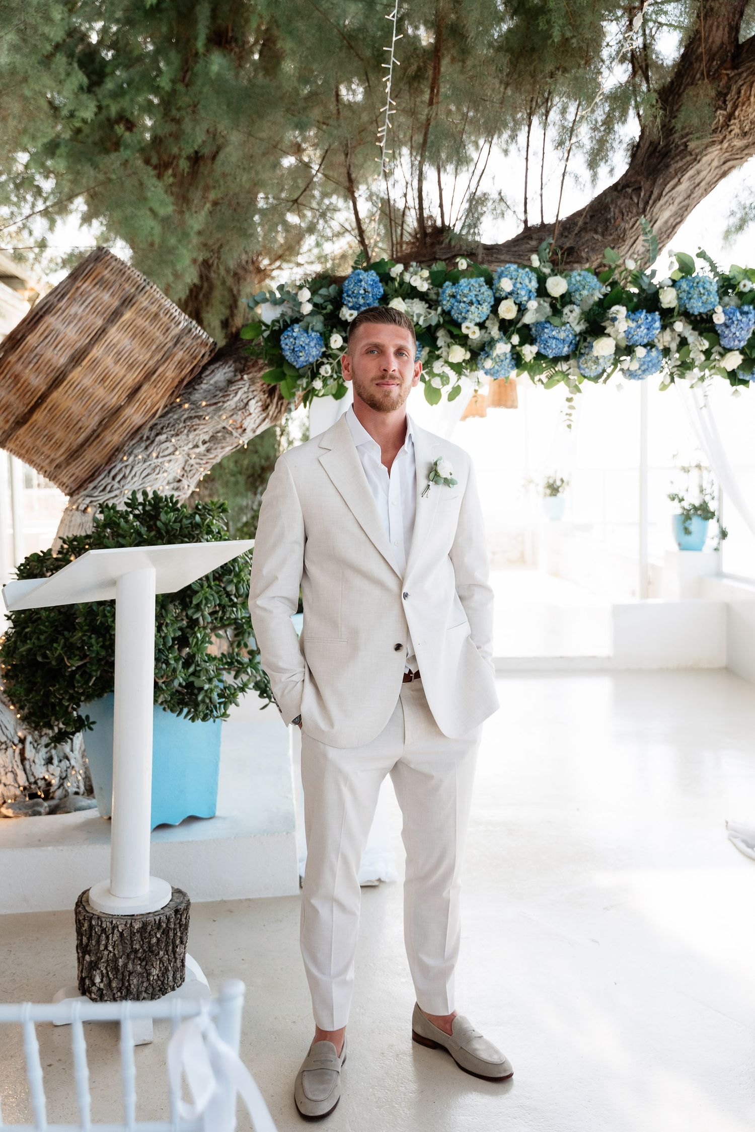 A groom in a light beige linen suit and brown shoes stands under a tree decorated with blue flowers at an outdoor wedding.