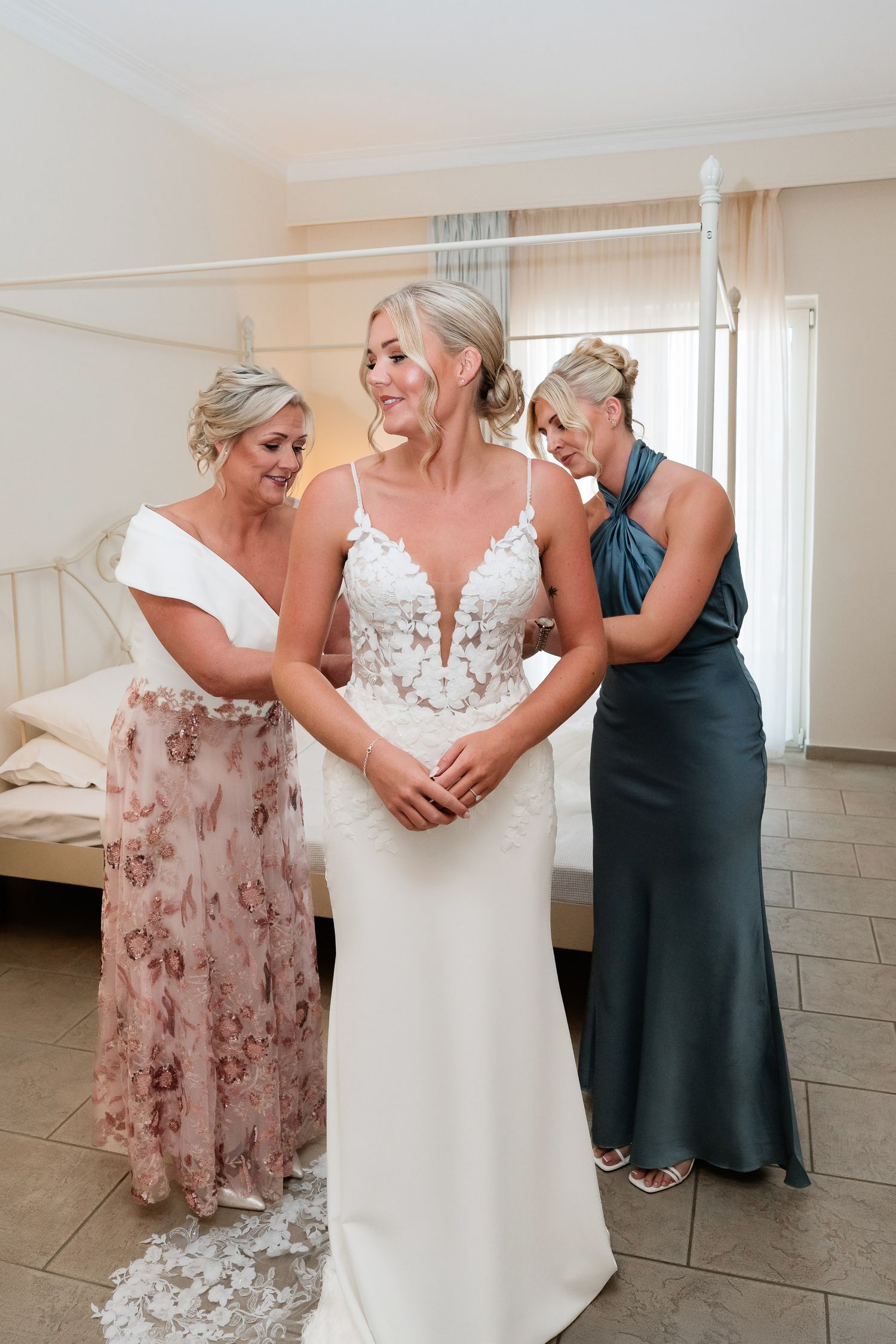 A tender moment during wedding preparations as bridesmaids in blush and navy help adjust the bride's white gown.